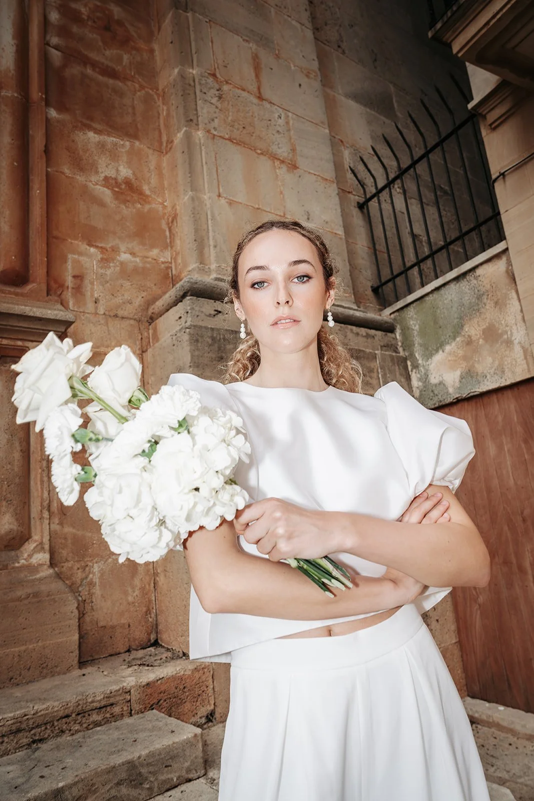 Junge Frau in weißem Kleid mit Blumenstrauß vor Backsteinmauer im Freien.