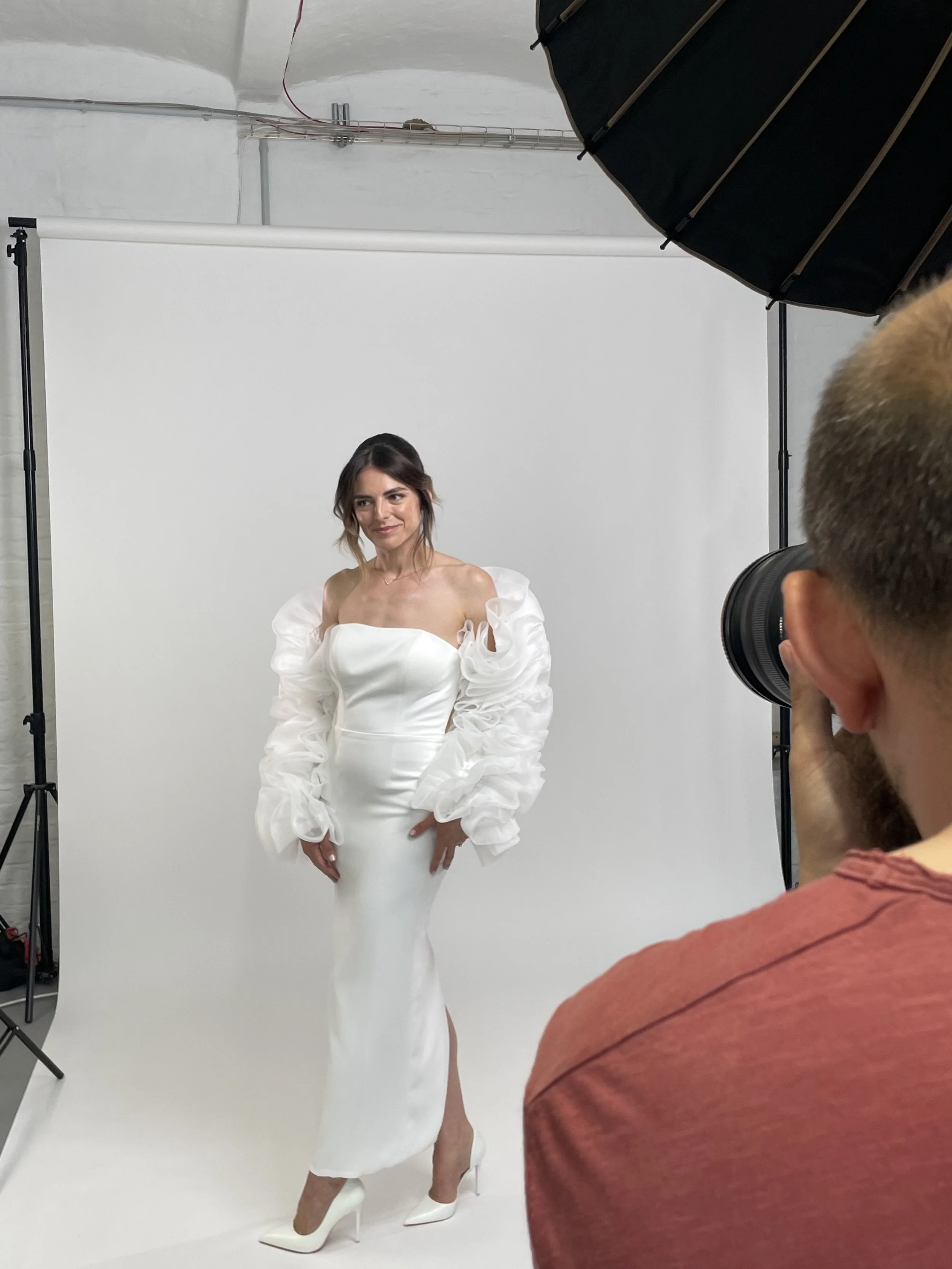 Frau in weißem Brautkleid bei Fotoshooting vor weißem Hintergrund, Fotograf fotografiert sie.