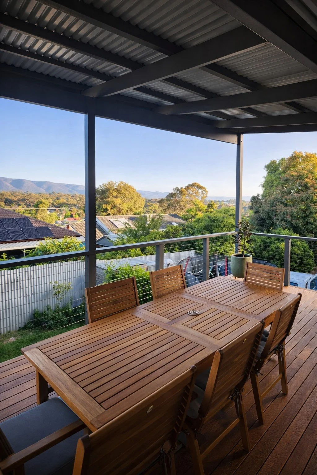 Outdoor patio with a wooden dining table and chairs under a metal roof, overlooking a suburban neighborhood and distant mountains, with trees and cars visible.