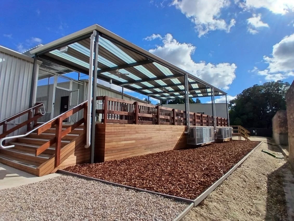 An outdoor structure with a metal roof, wooden stairs with handrails, and a wooden deck with railing. The deck has air conditioning units underneath and is set against a building with a metal exterior. The sky is partly cloudy with blue and white clouds visible.