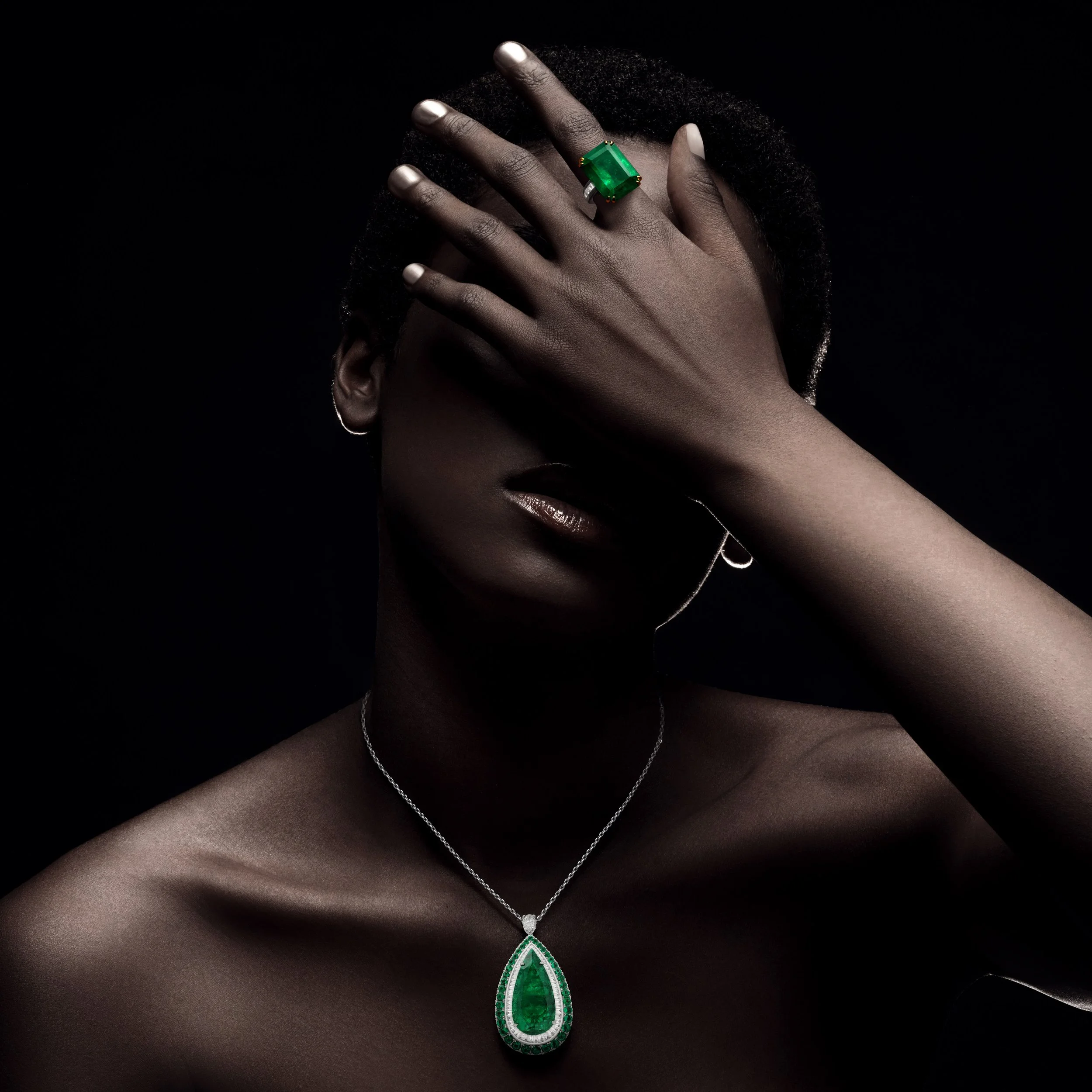 A person with dark curly hair covering their face with their hand, wearing a large emerald green ring and an emerald pendant necklace, set against a black background.