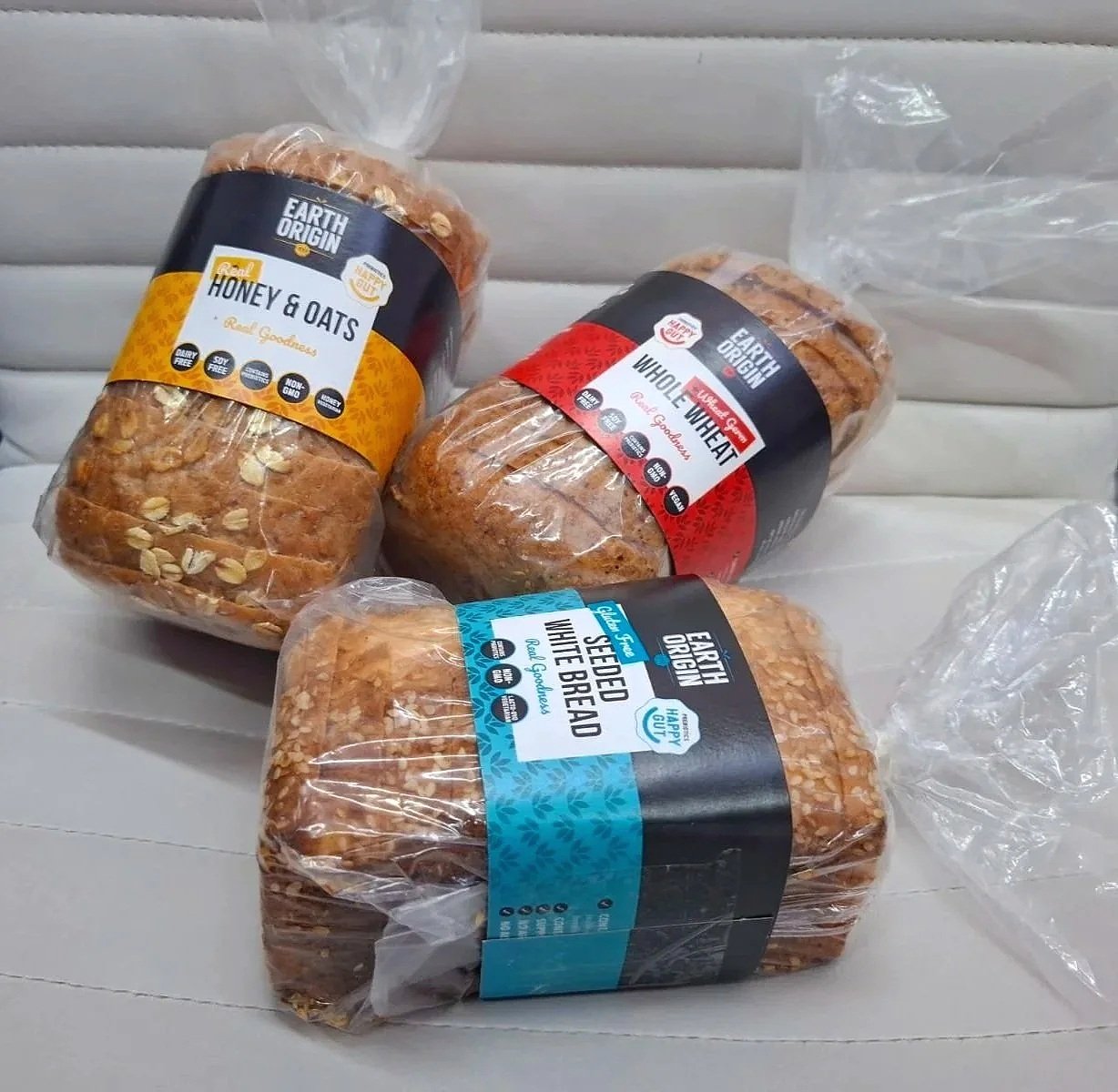 Three loaves of sliced bread from Earth Origin with different labels: Honey & Oats, Whole Wheat, and Seeded White Bread, on a white surface, with plastic wrap around the bread