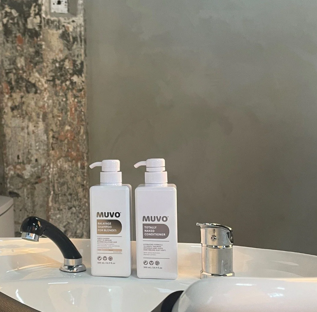 Two white bottles of hair care products labeled 'Muvo Balayage Shampoo for Blondes' and 'Muvo Totally Naked Conditioner' on a white sink with a silver faucet and a black handle, against a gray wall and exposed brick wall background.