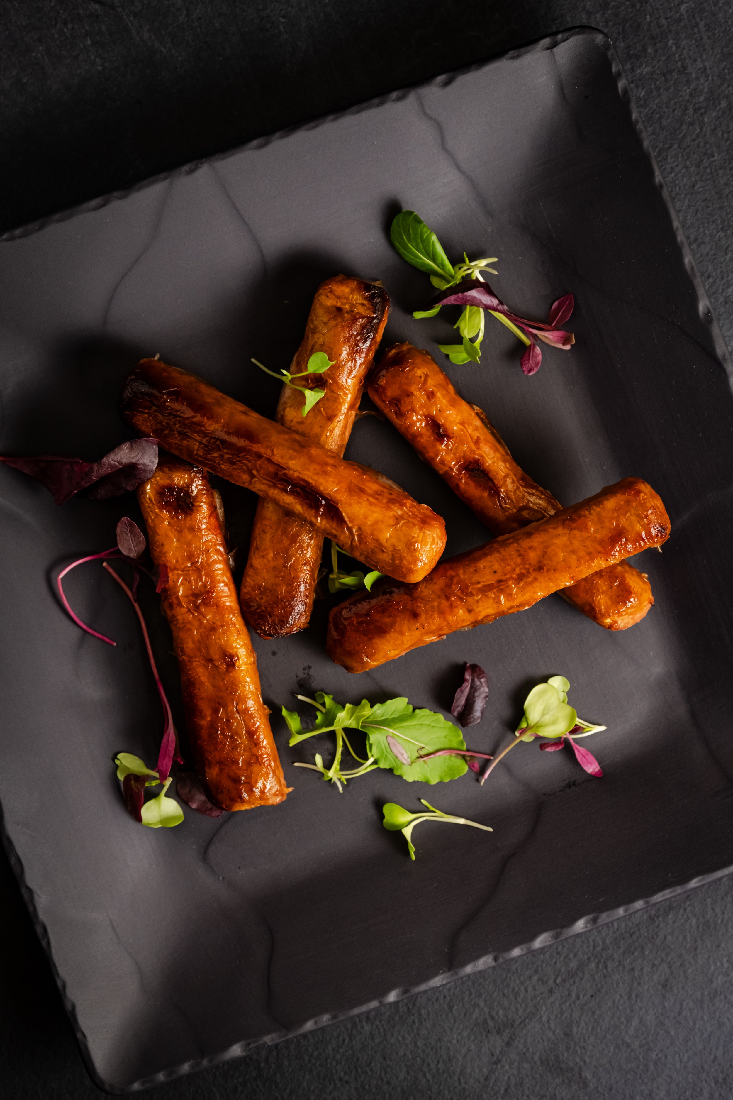 Six cooked sausages garnished with microgreens on a black square plate.