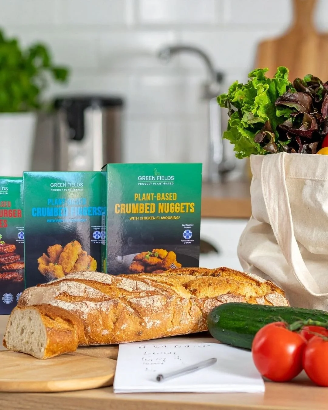 A loaf of bread, a cucumber, three tomatoes, plant-based crumbled nuggets, plant-based chicken-flavored nuggets, and a bag of leafy greens on a kitchen counter with packaging and a notepad.