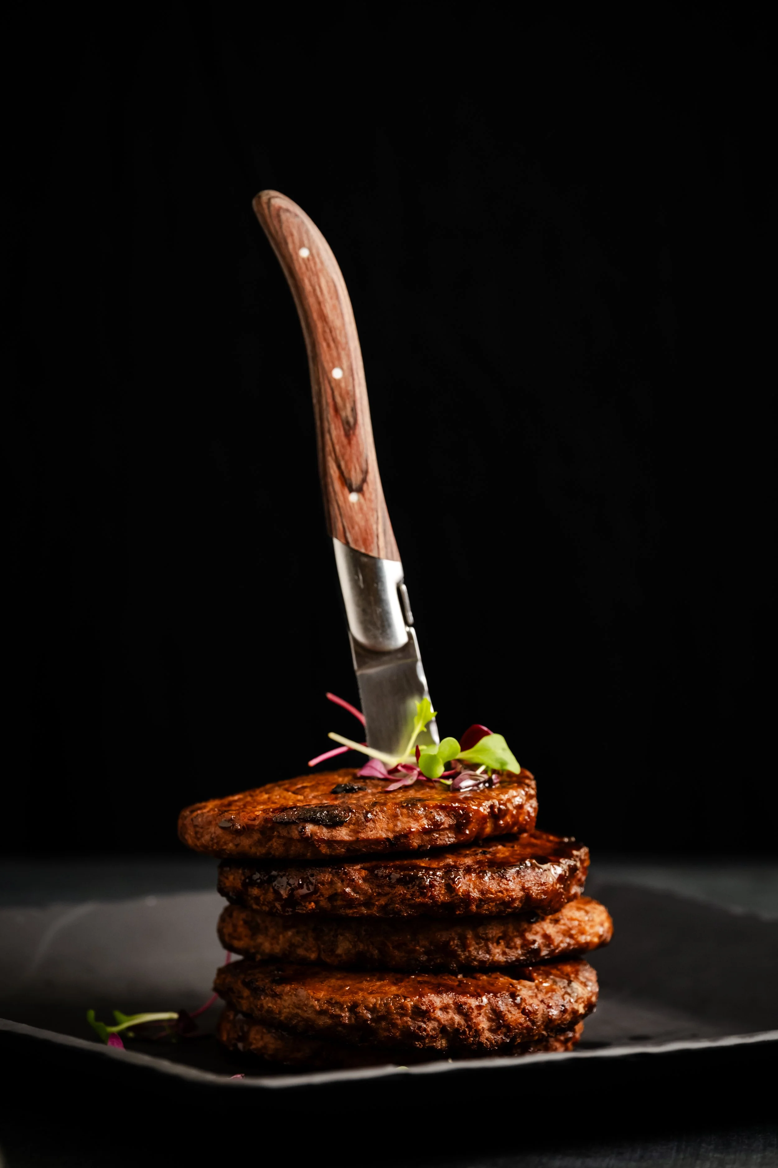 Stack of grilled burger patties with microgreens and a steak knife inserted into the top patty, dark background.