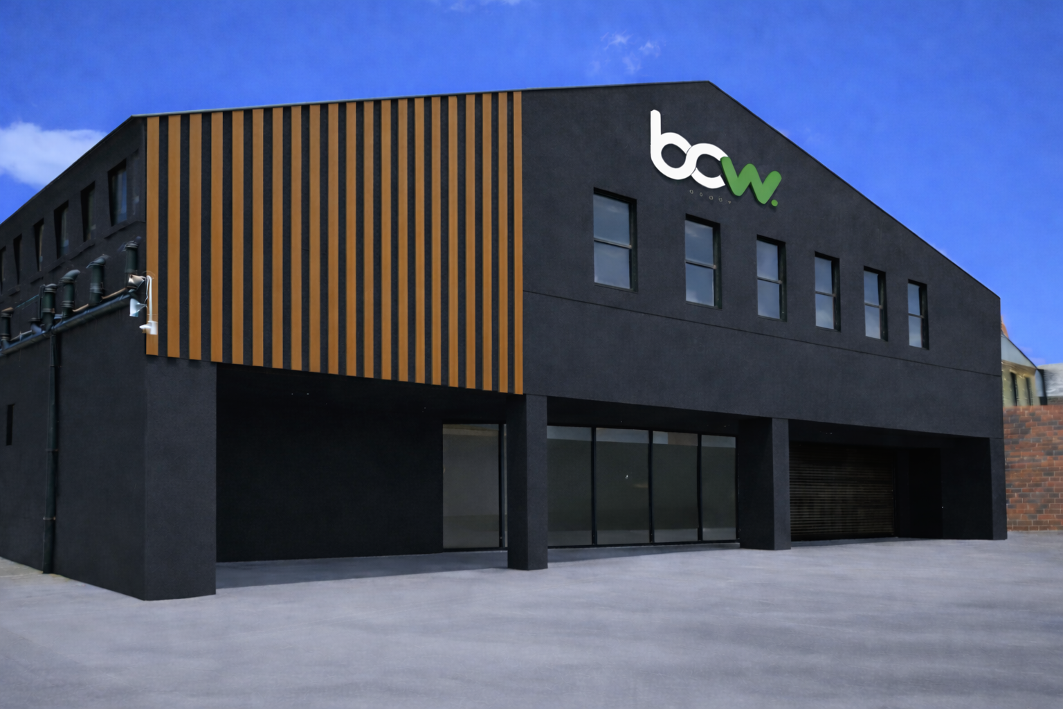 A modern, two-story black building with wooden slats on one side and large glass windows on the ground floor. A sign reading 'bcw' is on the upper corner. The sky is blue with some clouds.