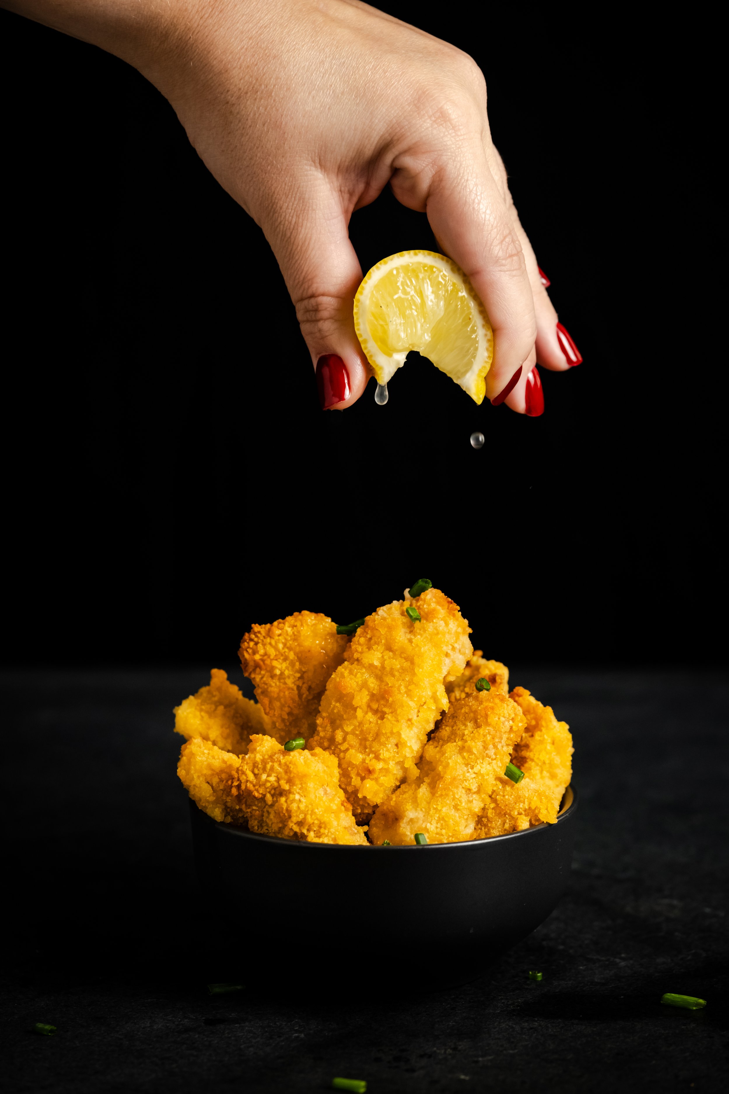 Hand squeezing lemon over a bowl of breaded fried chicken tenders garnished with chopped green onions, against a black background.