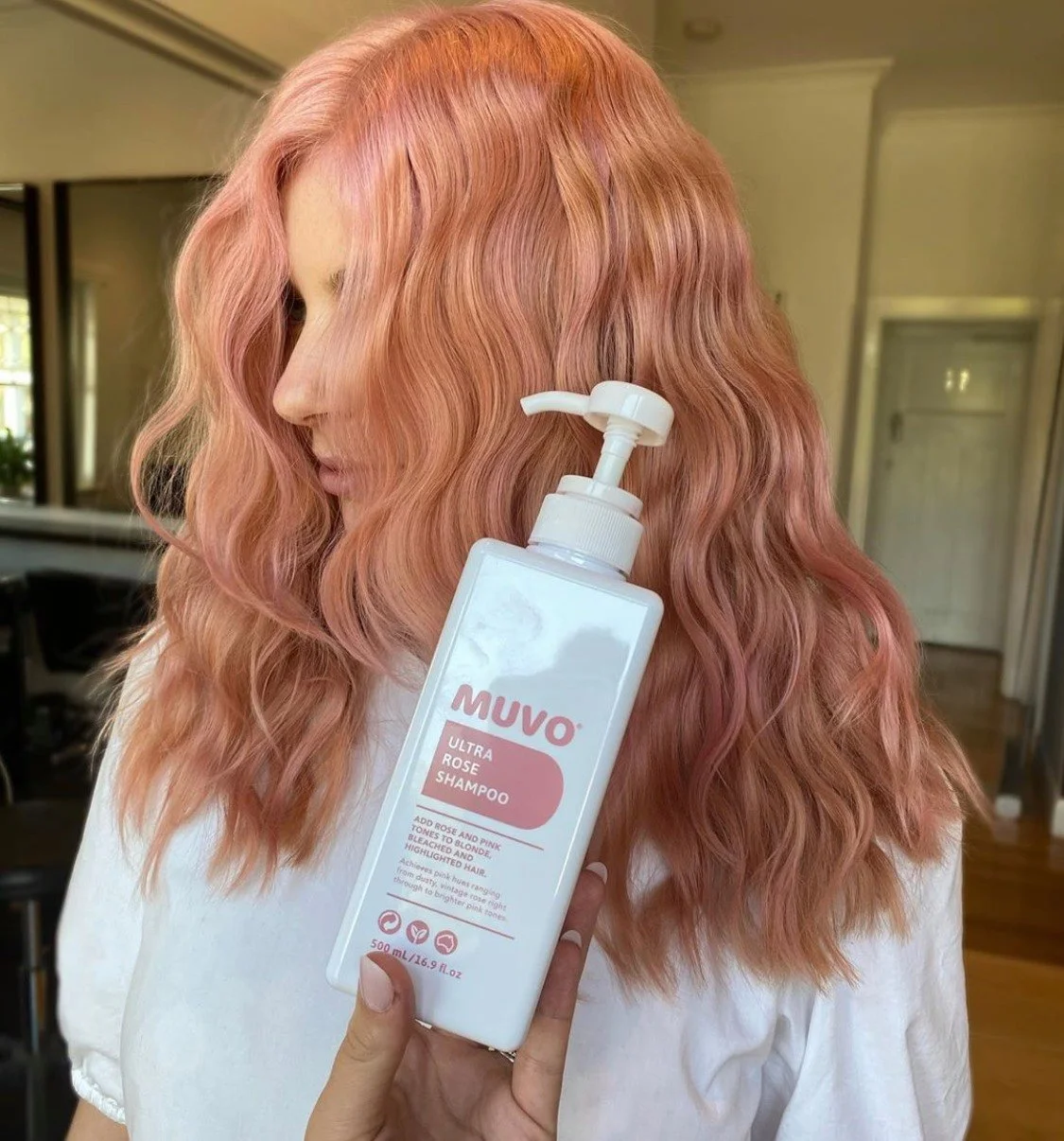 A woman with wavy pink hair holding a bottle of MUVO Ultra Rose Shampoo. The background shows a home interior with a mirror, a door, and some furniture.