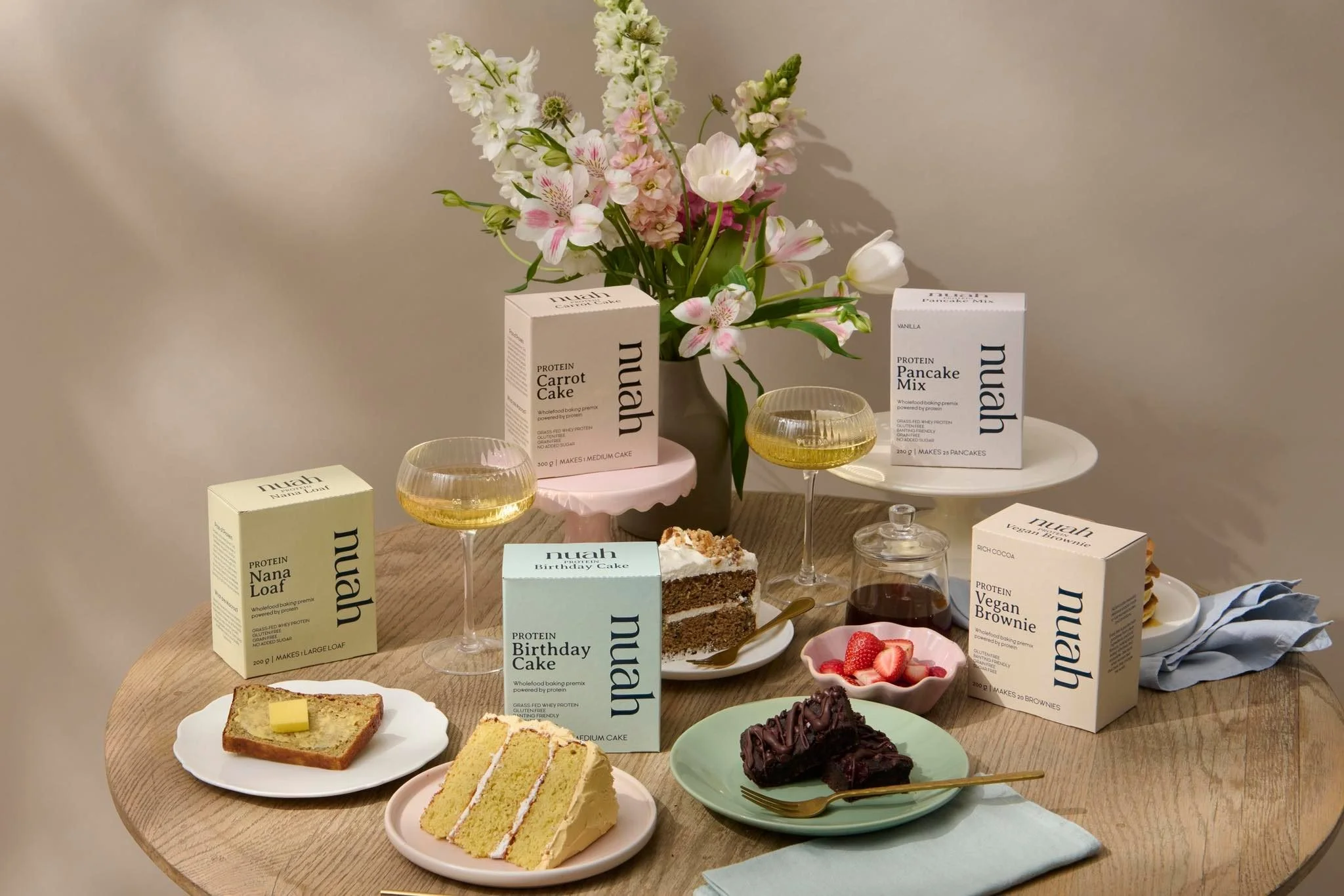 A variety of vegan cakes and snacks on a wooden table, with two wine glasses filled with a yellow beverage, and a floral vase with pink and white flowers at the center.