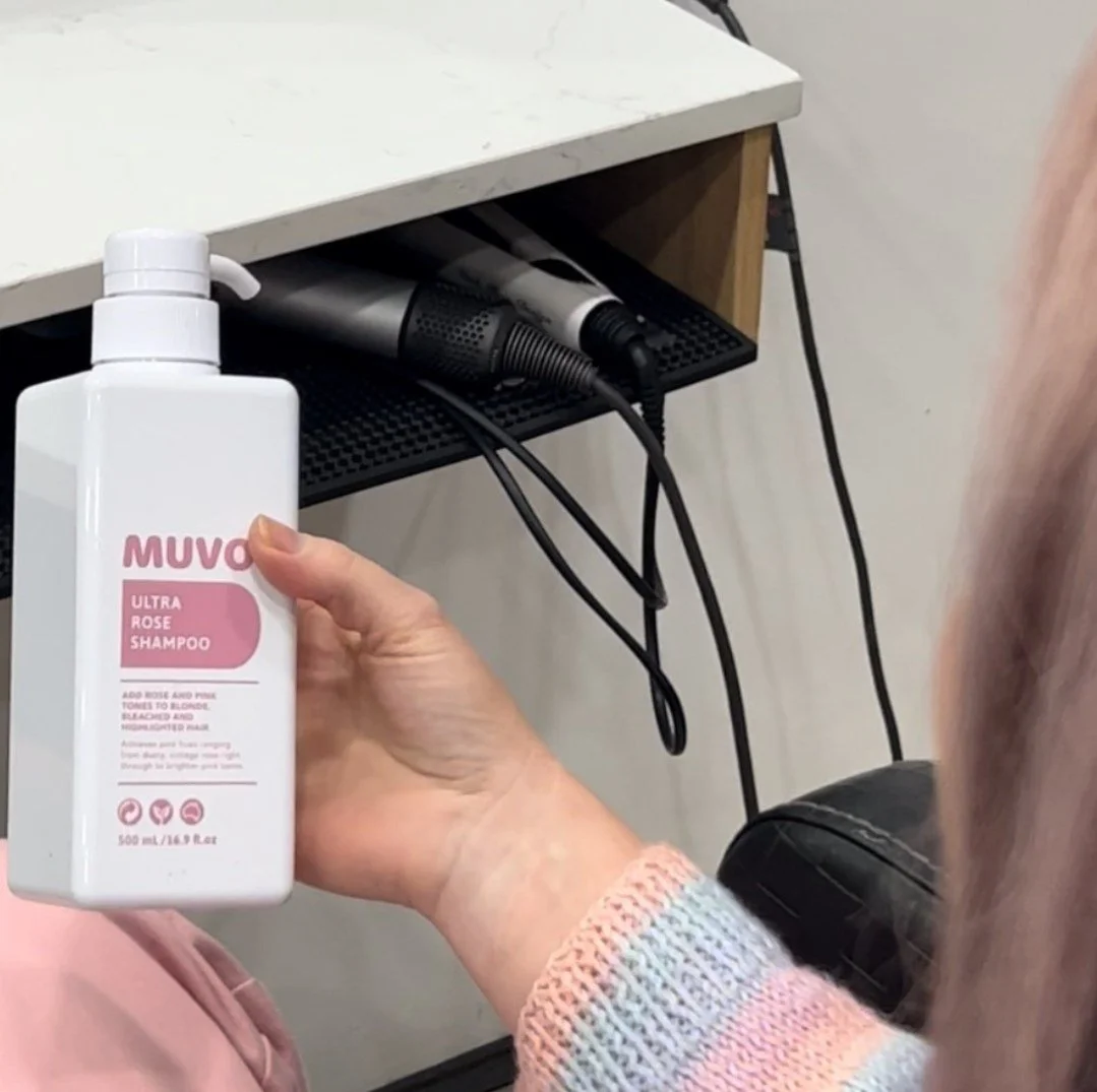 Person holding a white bottle of MUVO Ultra Rose Shampoo in a hair salon, with hair styling tools on a black shelf in the background.