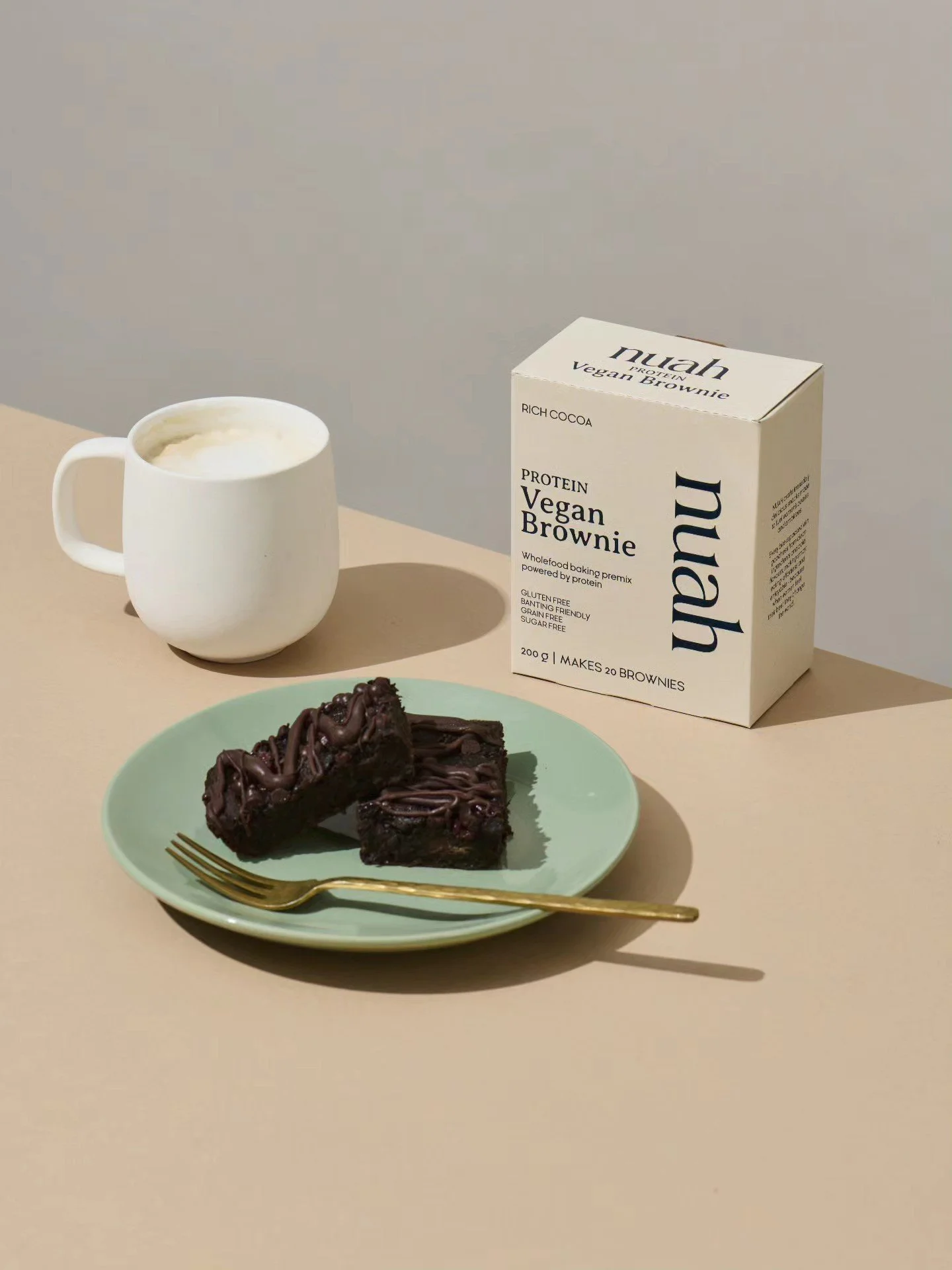 A plate with two pieces of vegan chocolate brownies, a gold fork, a white mug of frothy beverage, and a box of vegan brownie mix on a light-colored table