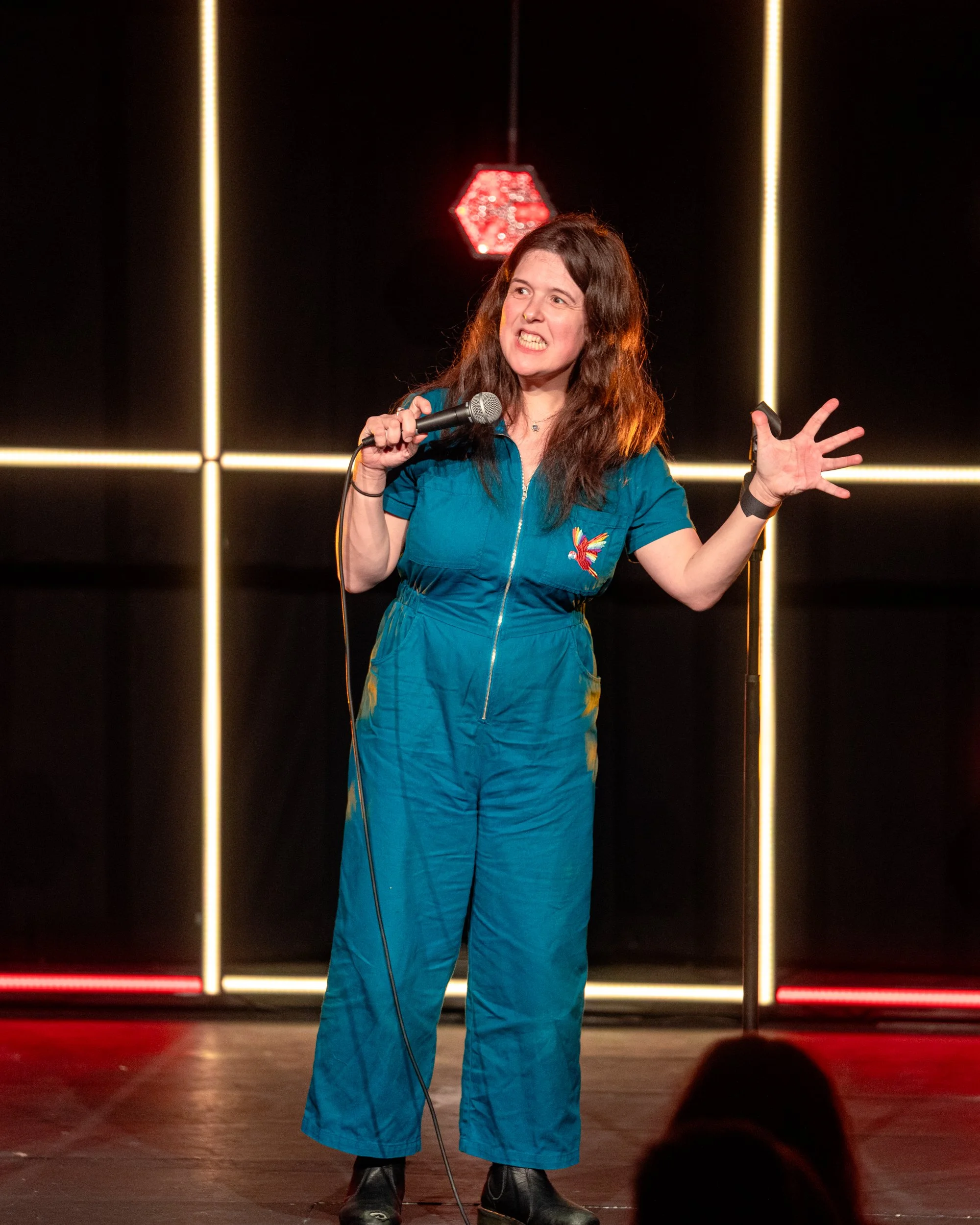 Rosie Jones performing stand-up comedy on stage, holding a microphone with her right hand, wearing a blue jumpsuit with a colorful bird embroidered on the chest, and making an expressive face with her left hand raised.
