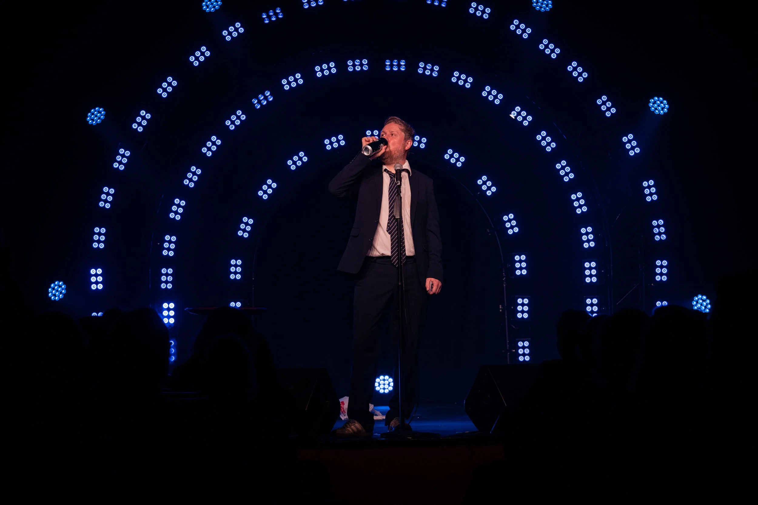 A man in a suit drinking from a can on stage with blue LED lights in an arc pattern behind him, performing in front of an audience.