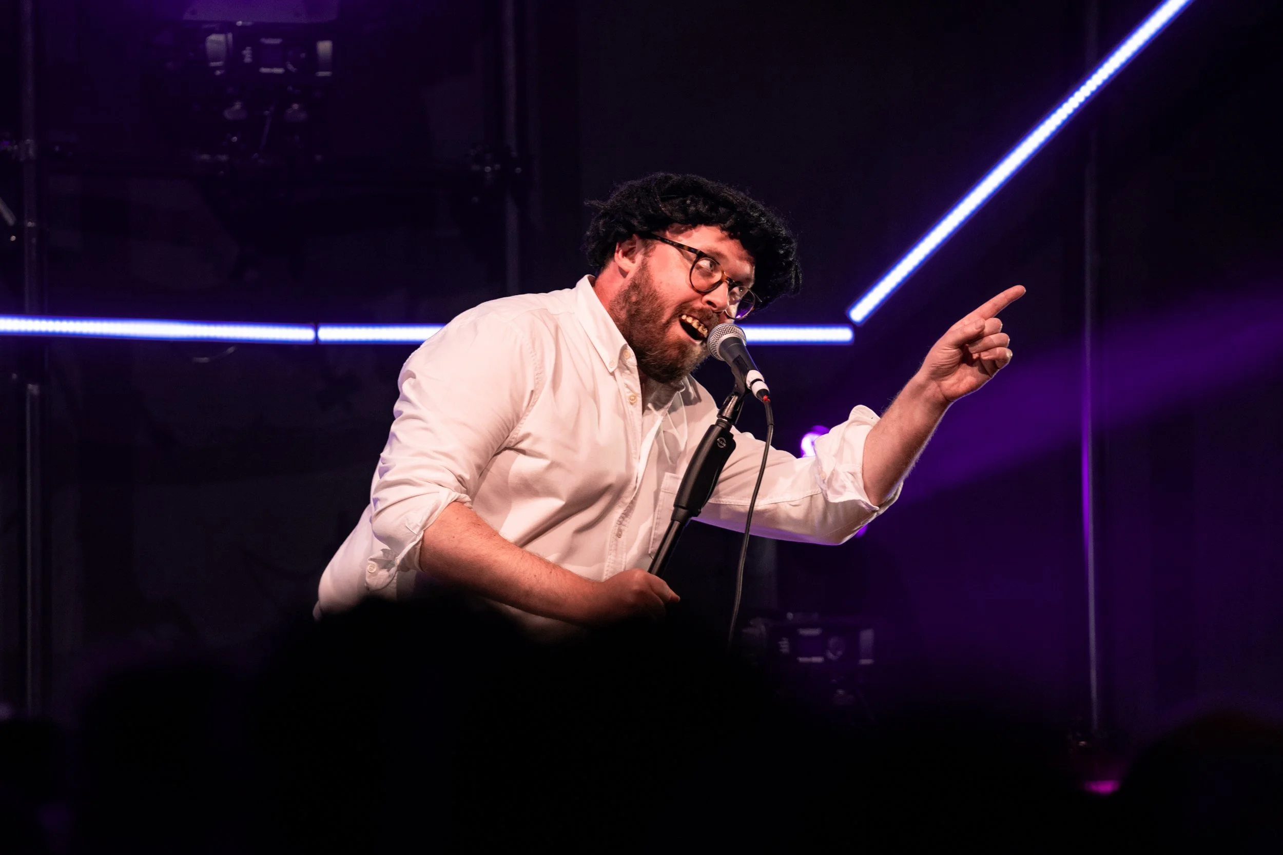 A man with curly hair, beard, glasses, wearing a white shirt, passionately singing into a microphone on stage with purple and blue lighting.