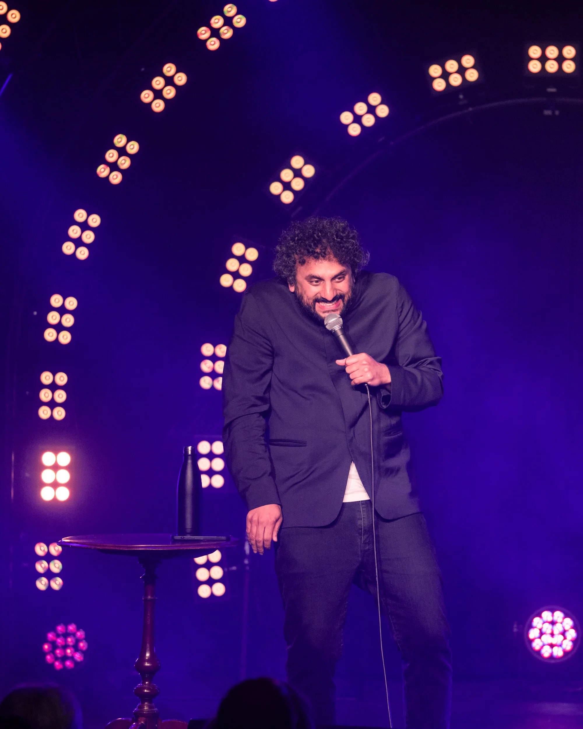 Comedian Nish Kuma - a man with curly hair and a beard - performs stand-up comedy on stage, holding a microphone. There is a small round table with a water bottle beside him. The stage is lit with pink and purple lights.