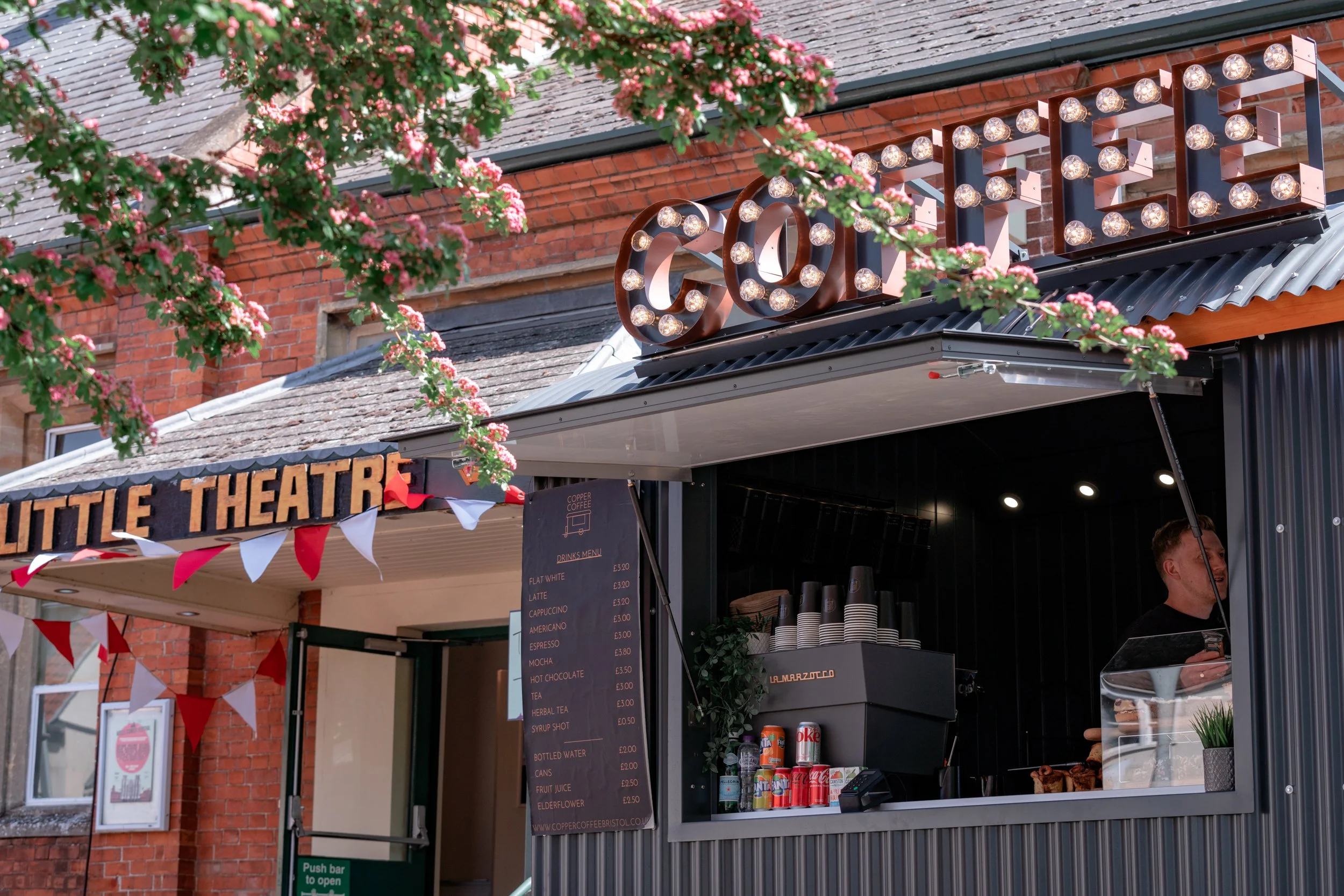 A small outdoor coffee stand named 'Copper Coffee' with a black exterior, a menu board, and a person serving coffee inside. There is a decorative sign with the word 'COFFEE' outlined with light bulbs and pink blossoms hanging above.