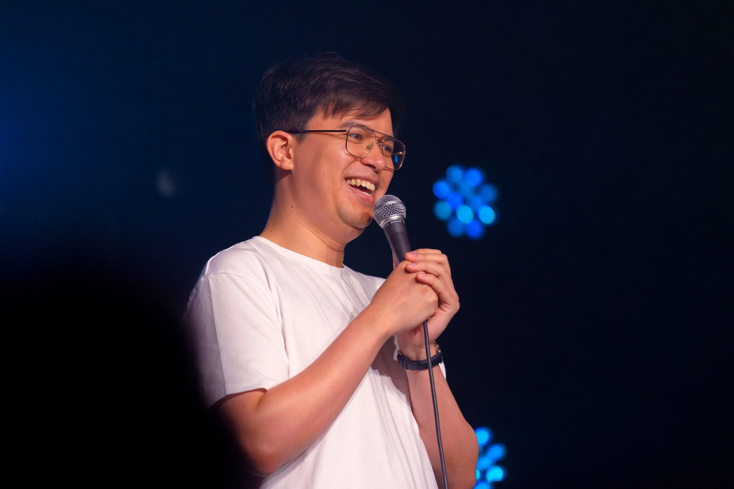 Phil Wang - a young man with glasses and short dark hair, wearing a white t-shirt - is smiling while holding a microphone and speaking on stage.