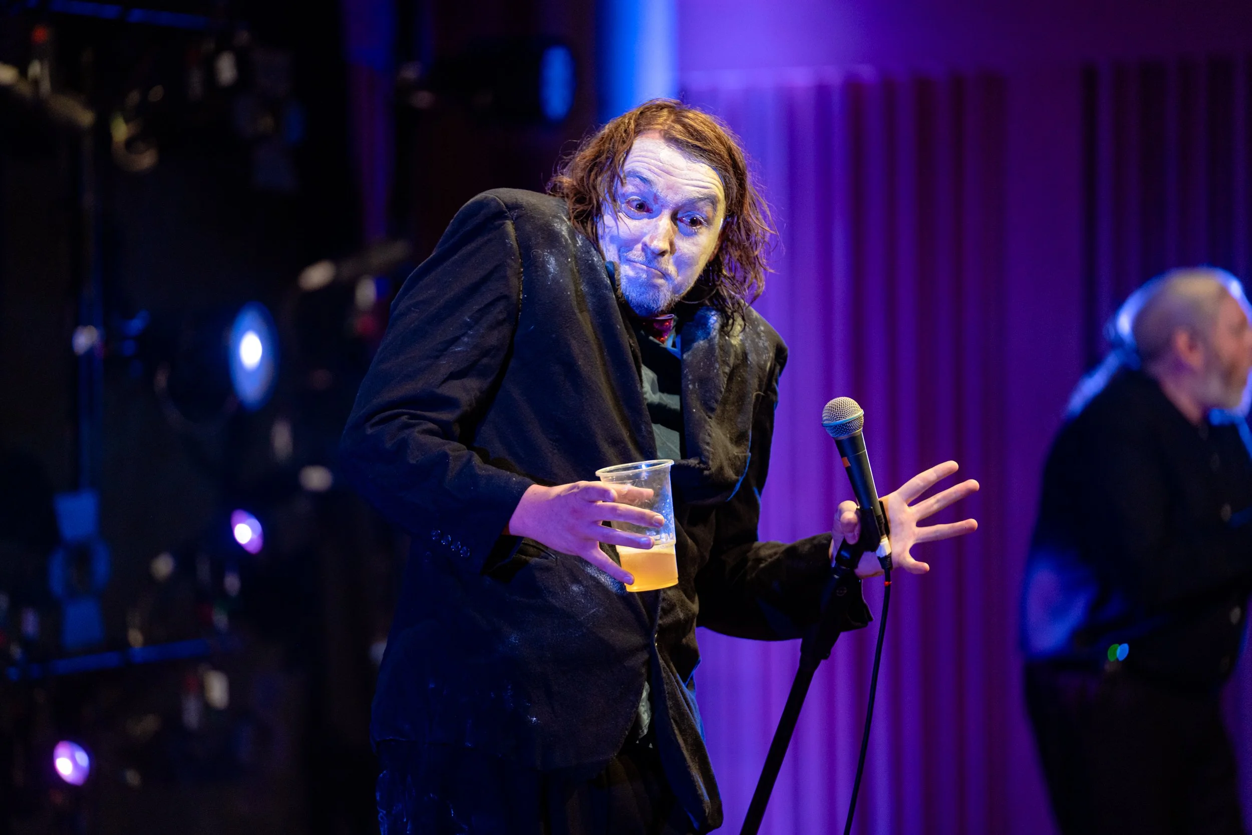 A man performing on stage, holding a drink in one hand and a microphone in the other, with purple stage lighting and a curtain in the background.