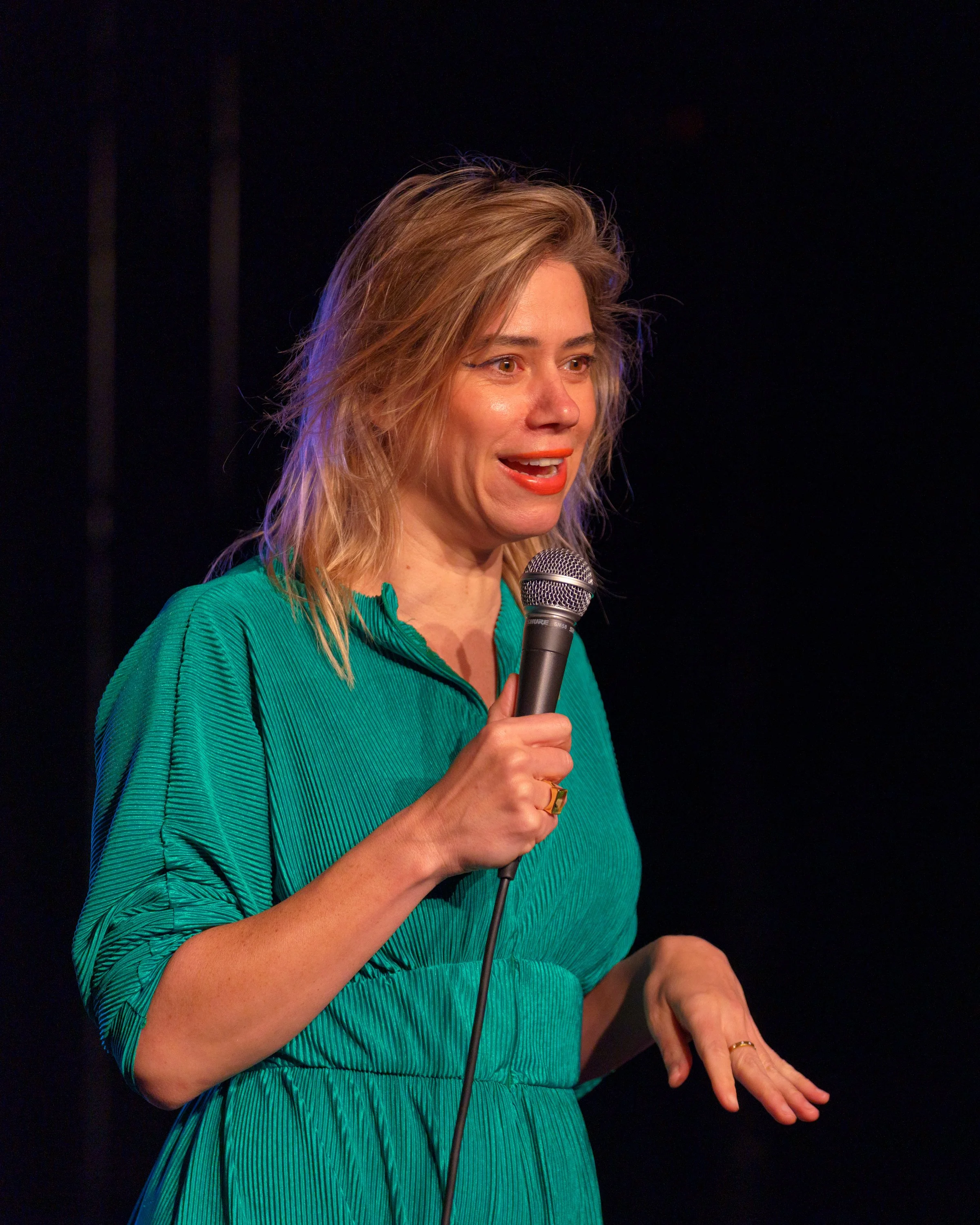 A woman in a teal dress holding a microphone and speaking on stage.