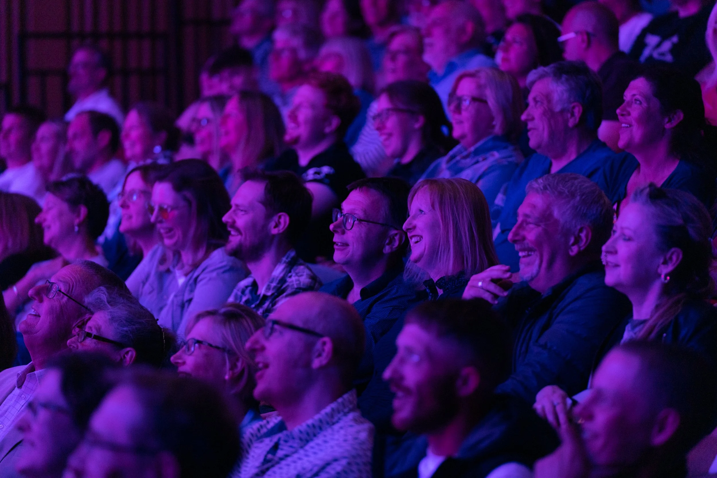 Audience of diverse adults laughing and enjoying a live performance or presentation in a dimly lit venue with purple lighting.
