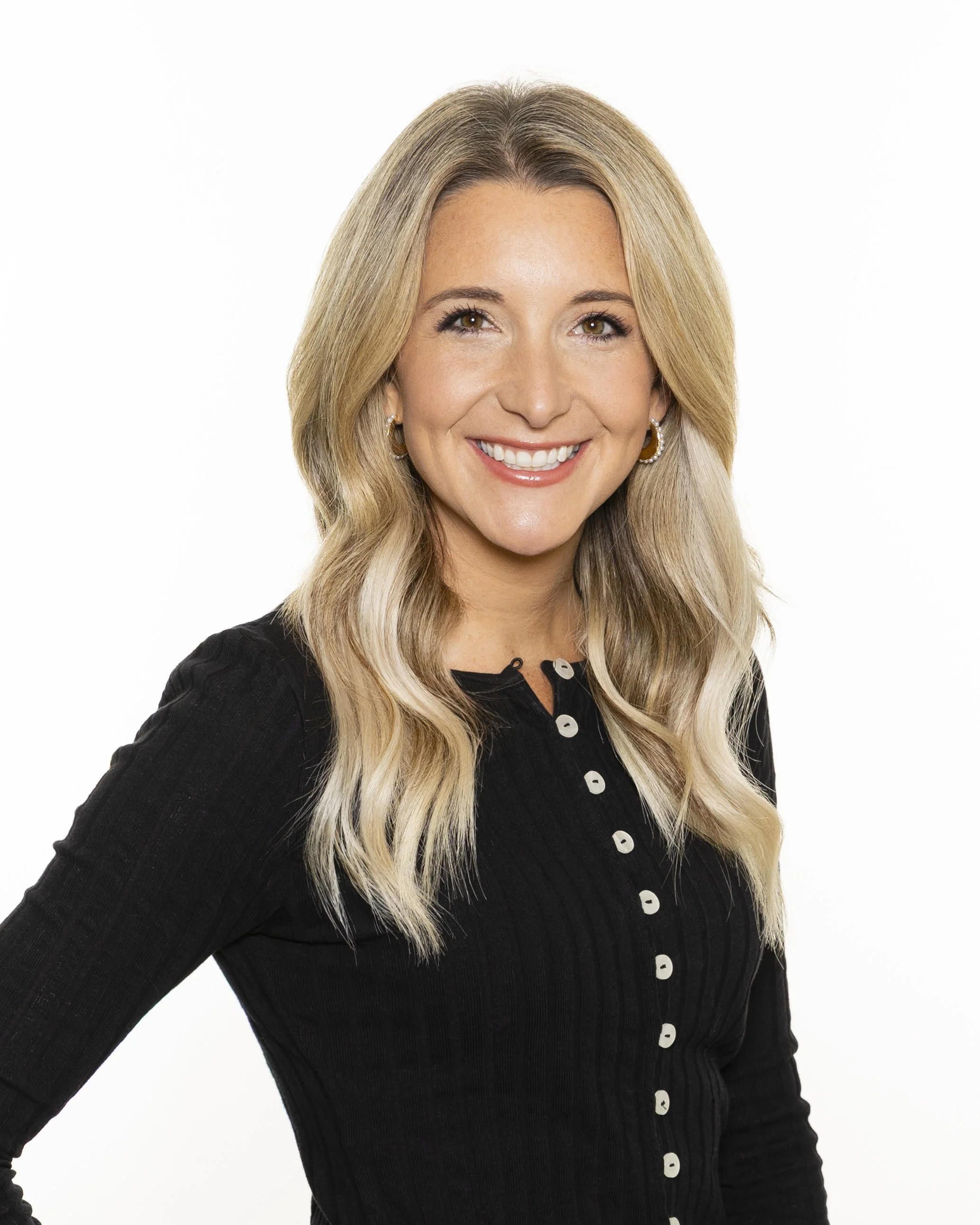 A woman with shoulder-length brown hair and blonde highlights, smiling, wearing a black blazer, against a plain white background.