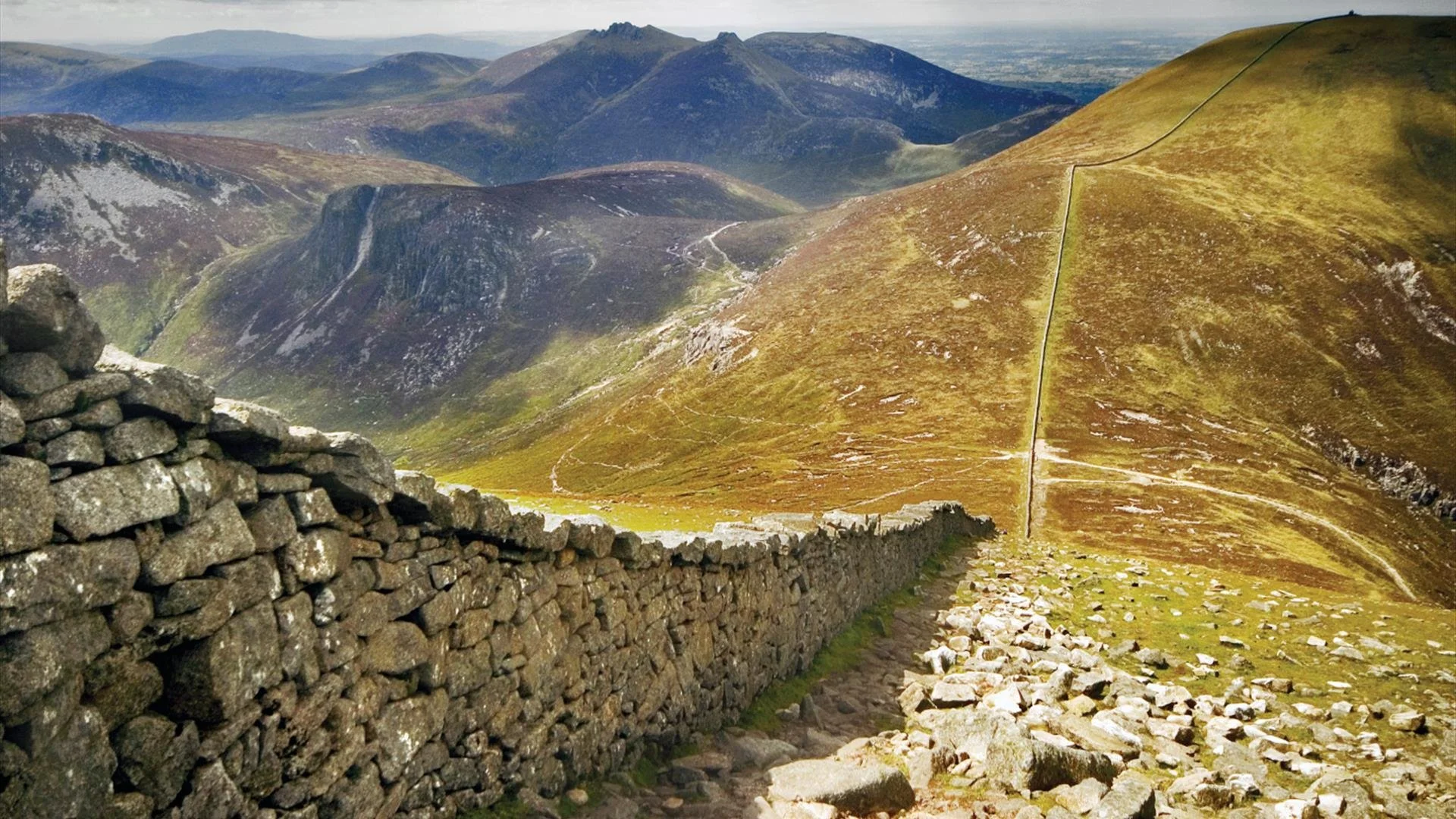 Mountain landscape with a stone wall in the foreground and rolling hills in the background.