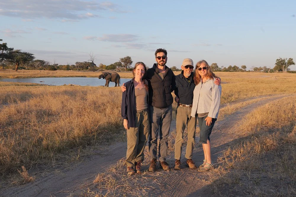 Four people standing together on a dirt path in a grassland with an elephant near a water body in the background, under a partly cloudy sky during sunset.