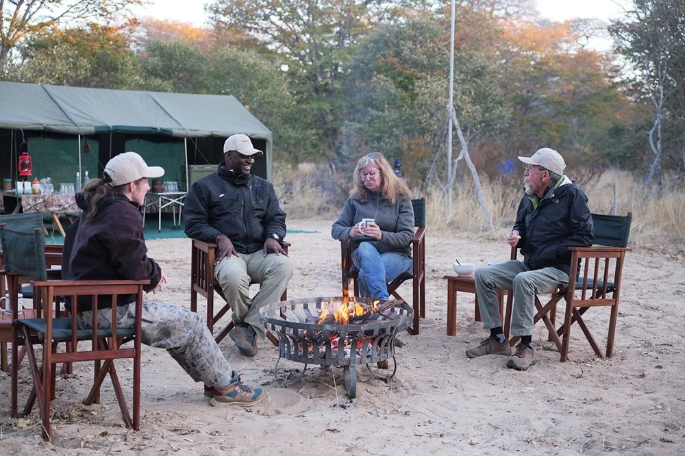 Four people sitting around a campfire on sandy ground, with a large green tent and trees in the background.