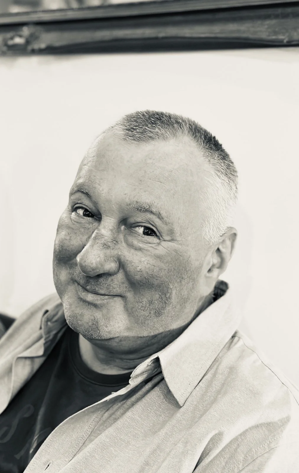 Black and white photo of a man with short hair, wearing a light button-up shirt over a dark T-shirt, smiling subtly.