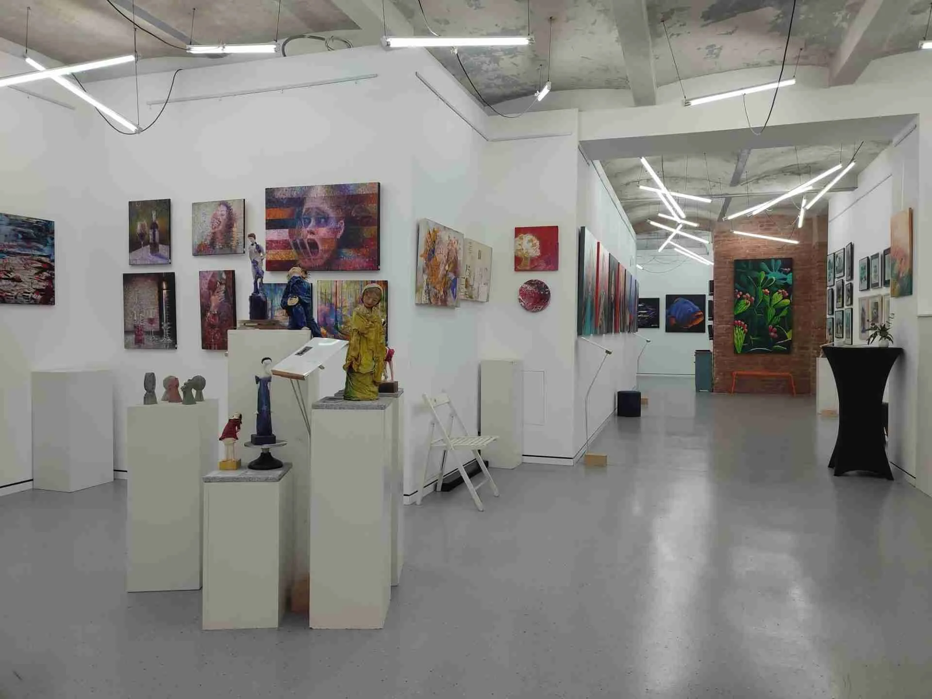 Interior view of an art gallery with various paintings and sculptures displayed on white walls and pedestals, featuring modern lighting fixtures and a brick wall section.