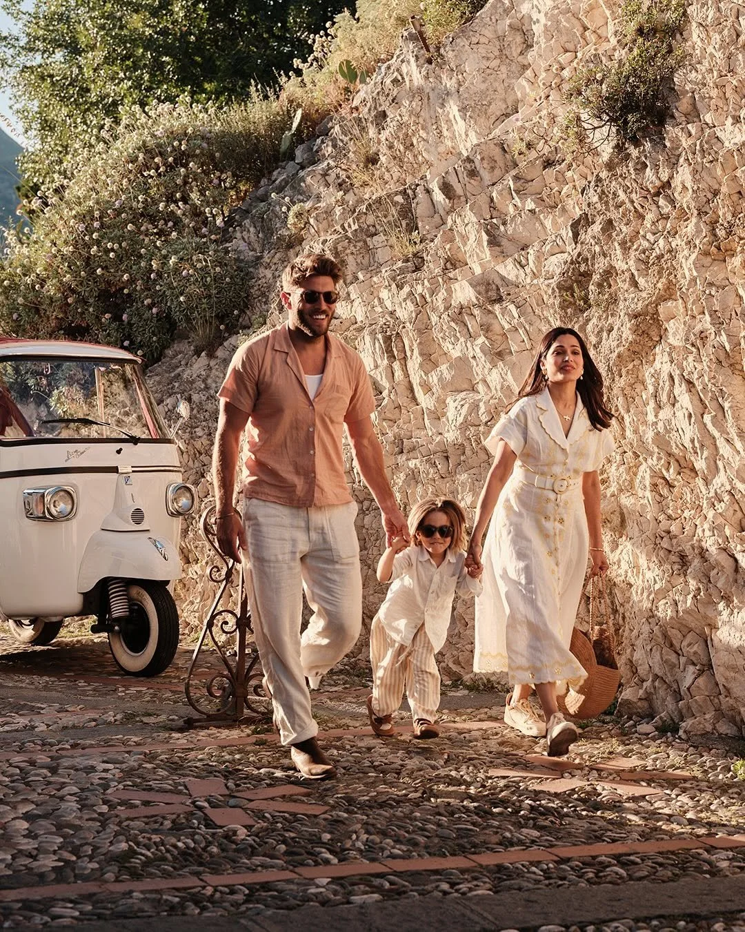 A family of four walking hand-in-hand outdoors on a sunny day, with rocky terrain and flowering bushes in the background, next to a vintage white scooter.