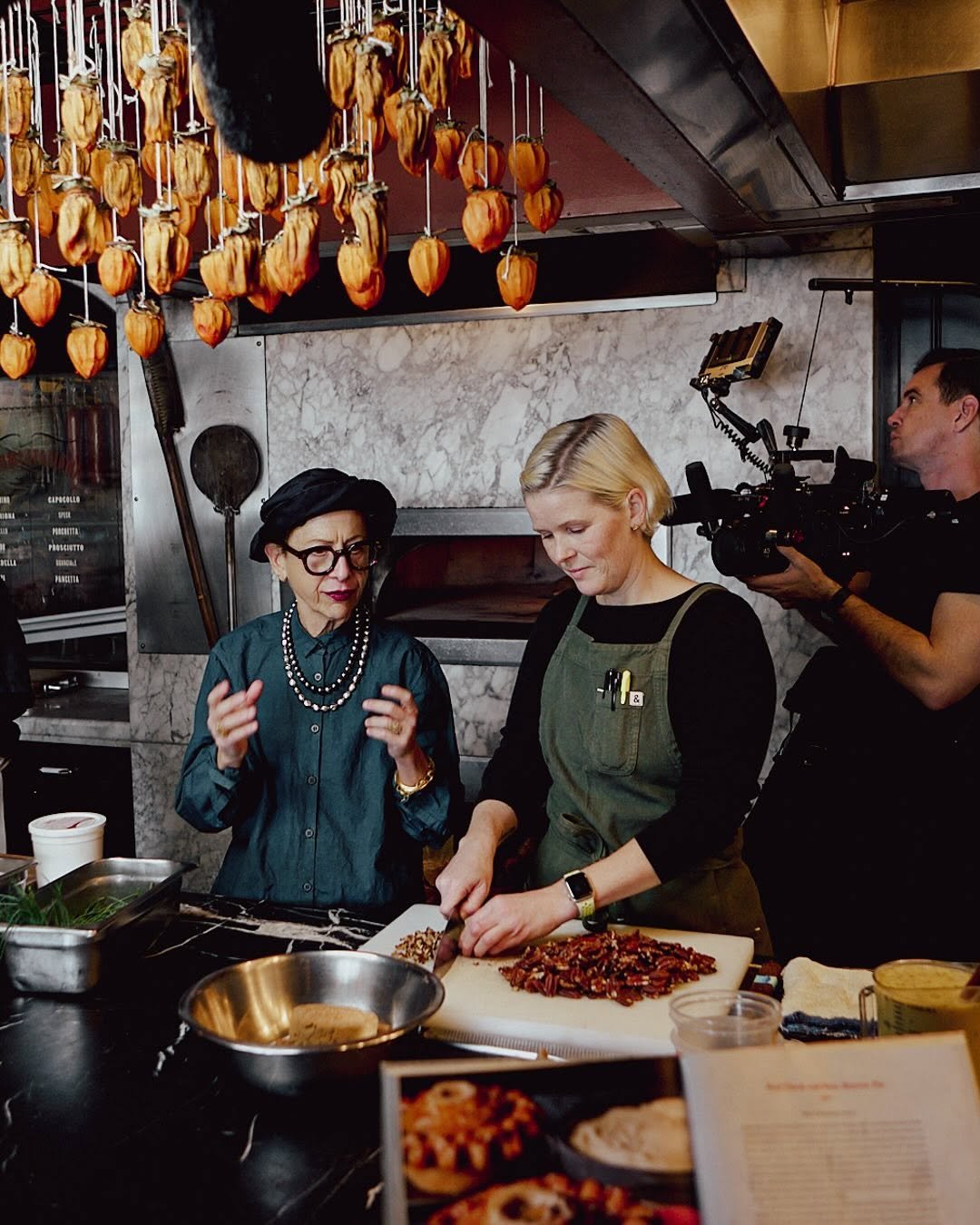 A woman in a black headscarf and glasses explains to a blonde woman wearing a green apron as she chops cooked meat in a kitchen, with a camera operator filming them. Dried tomatoes hang from the ceiling above, and a menu is visible in the background.