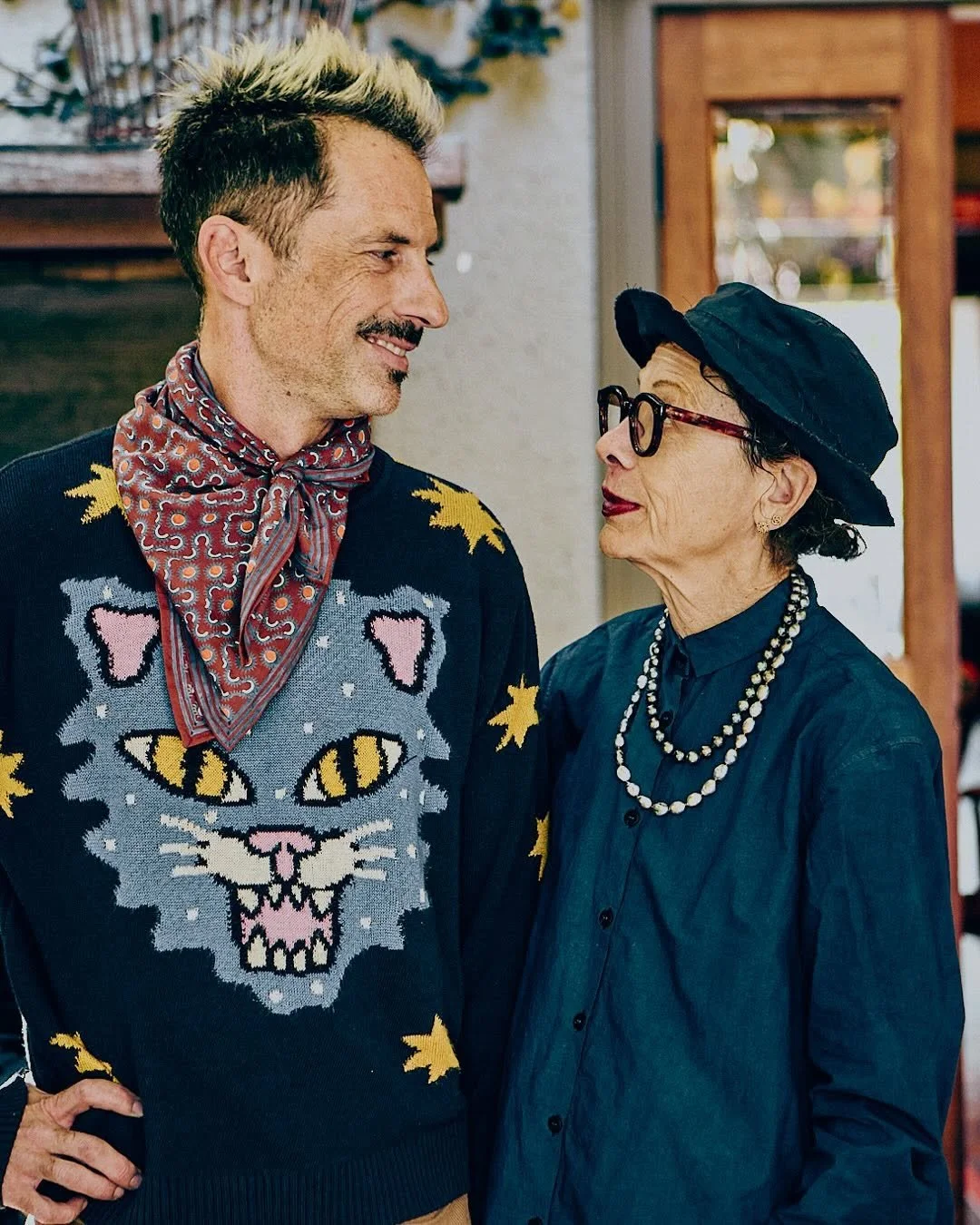 A young man with a styled, blonde-more-than-blonde hairdo and a mustache wearing a dark sweater with colorful cat face and star patterns, paired with a tie or scarf. An older woman with dark hair, glasses, a navy hat, a dark blue shirt, and a pearl necklace looks at him affectionately in a warm indoor setting.