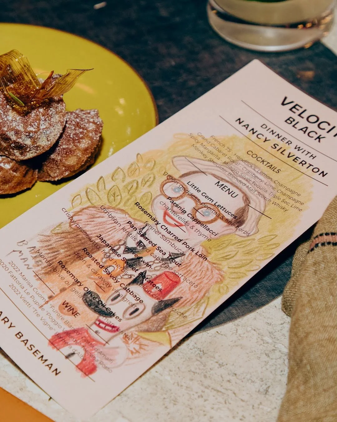 A colorful illustrated menu and a dessert on a bright green plate. The menu features a cartoon character and lists various food and cocktail options. The dessert appears to be topped with a piece of caramelized candied fruit.