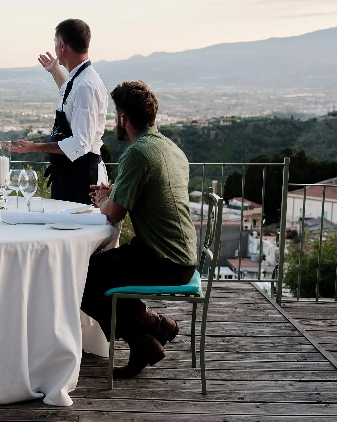 A man sitting at an outdoor dining table on a balcony, looking at a waiter who is speaking, with a scenic view of mountains and a valley in the background during sunset.