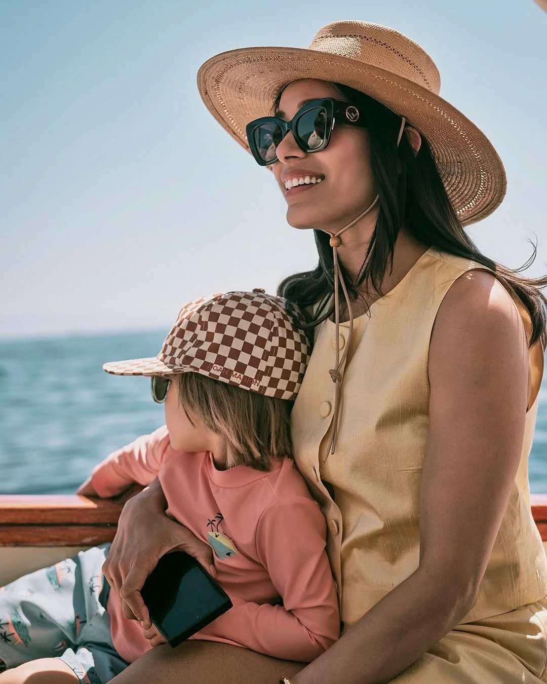 A woman and a young girl sitting on a boat near the water, enjoying a sunny day. The woman is wearing a wide-brimmed hat and large sunglasses, while the girl is wearing a checkered cap and holding a phone.