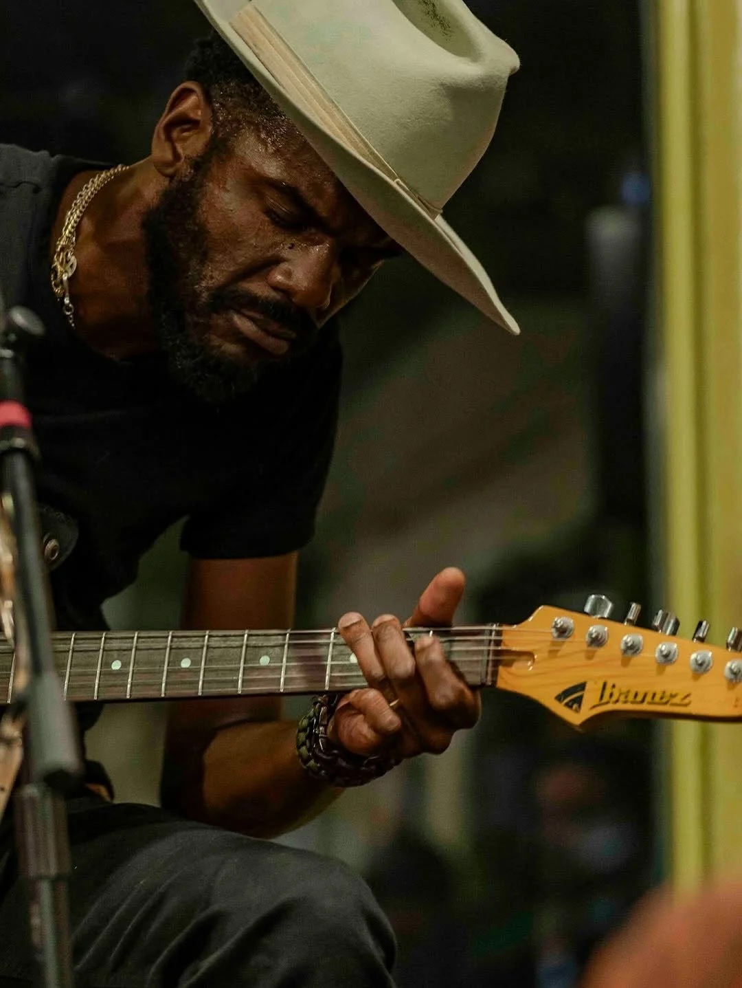 A man wearing a wide-brimmed hat and a gold chain necklace playing an acoustic guitar.