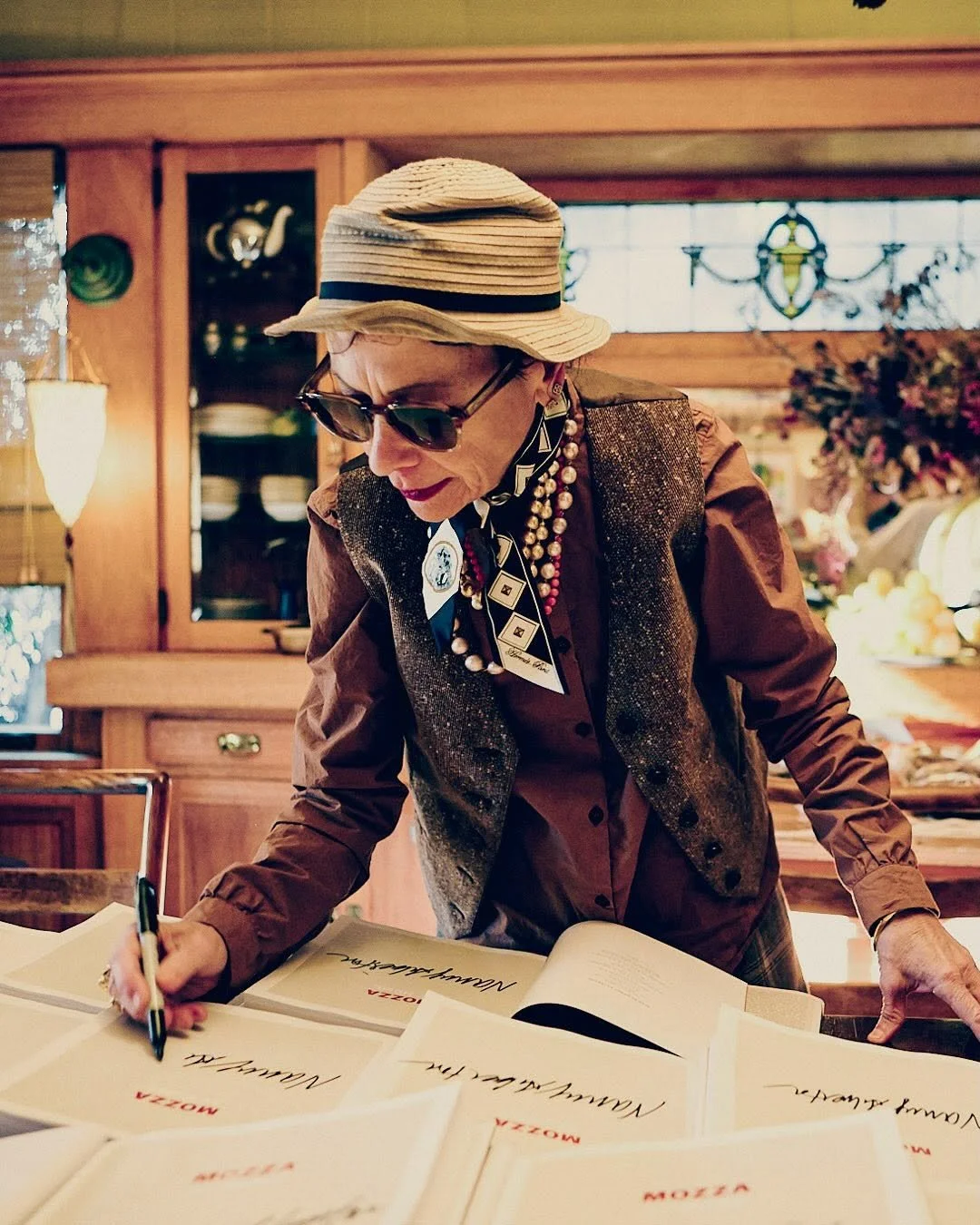 An elderly woman with sunglasses, a hat, and jewelry signs multiple menus on a table in a cozy, wooden interior.