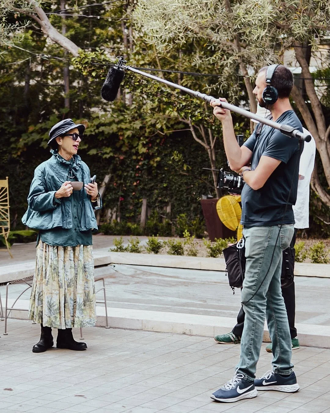 A woman holds a cup and phone while being interviewed by a man with a boom microphone. The scene is outdoors with trees and plants in the background.