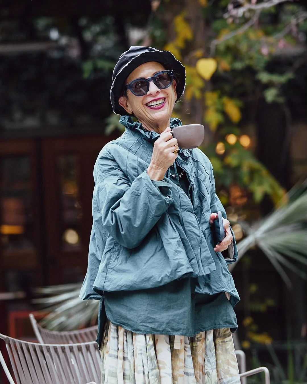 An elderly woman wearing sunglasses, a black hat, and a blue jacket, smiling while holding a cup outdoors.