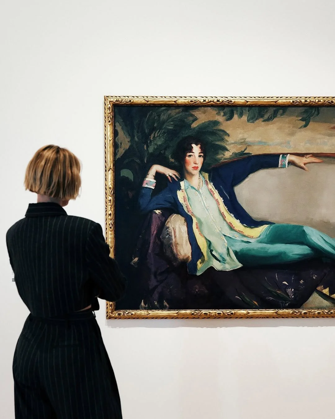 A woman with short blonde hair, dressed in a black pinstripe suit, is looking at a large framed portrait of a woman reclining with one arm outstretched and the other resting on her hip, set against a colorful background.