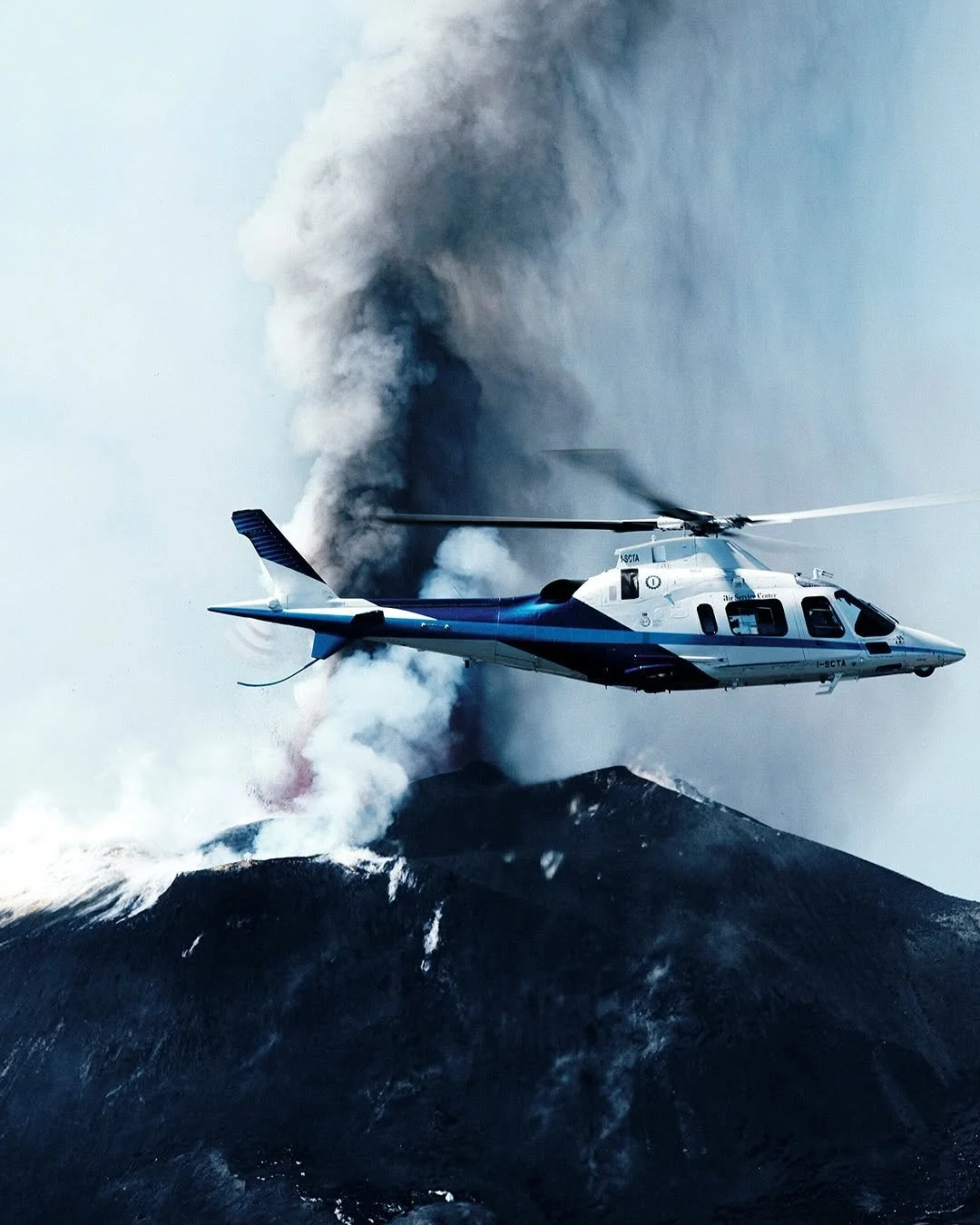 A helicopter flying over a volcanic eruption with smoke and ash billowing into the sky.