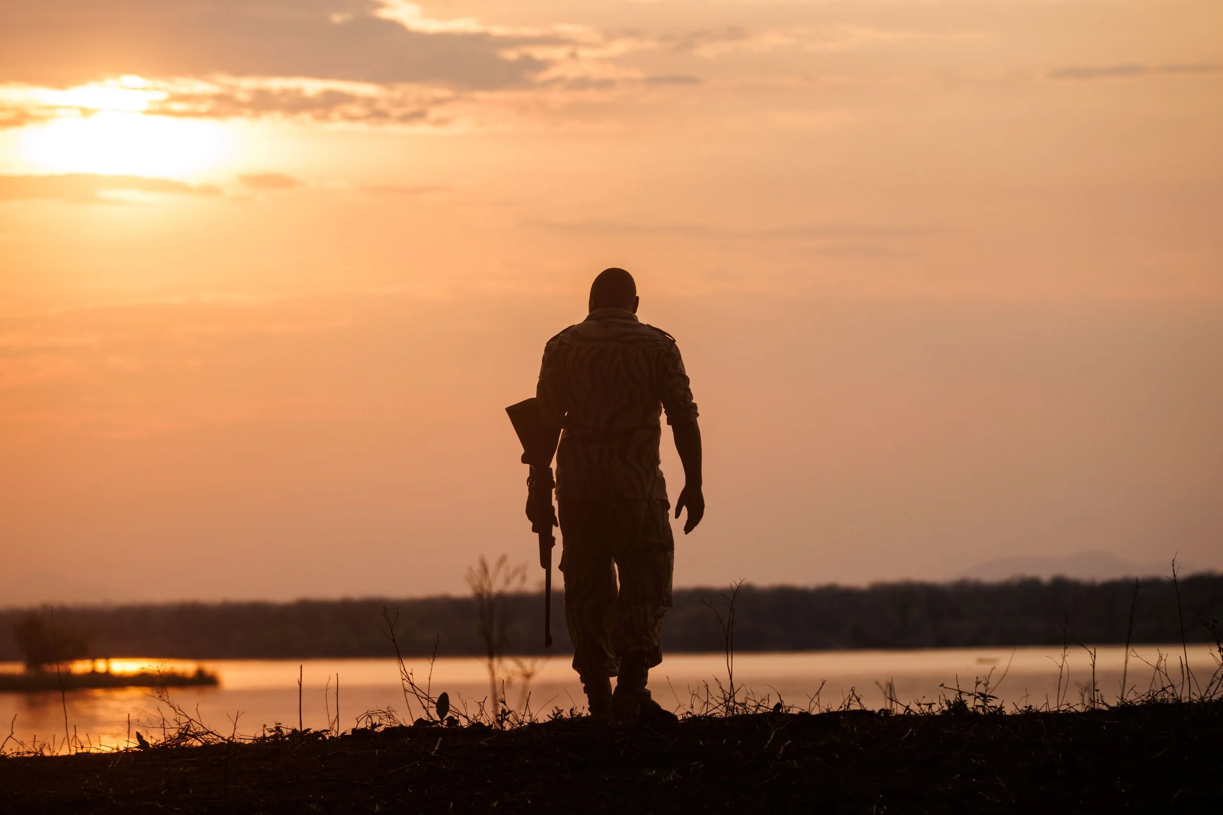Silhouette of a soldier walking along a shoreline during sunset, carrying a rifle.