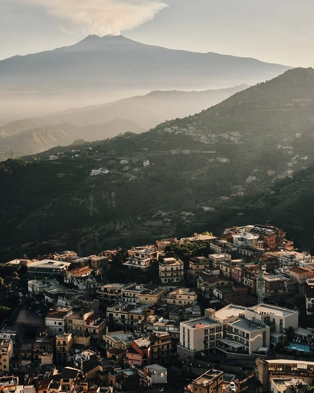 Urban neighborhood with multi-story buildings at the base of green, hilly mountains, with a mountain in the background emitting a small cloud of smoke or steam.