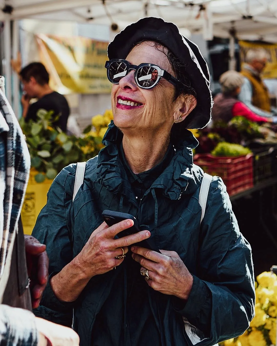 An elderly woman with short gray hair wearing black sunglasses, a black hat, a dark jacket, and a backpack, smiling at a market.