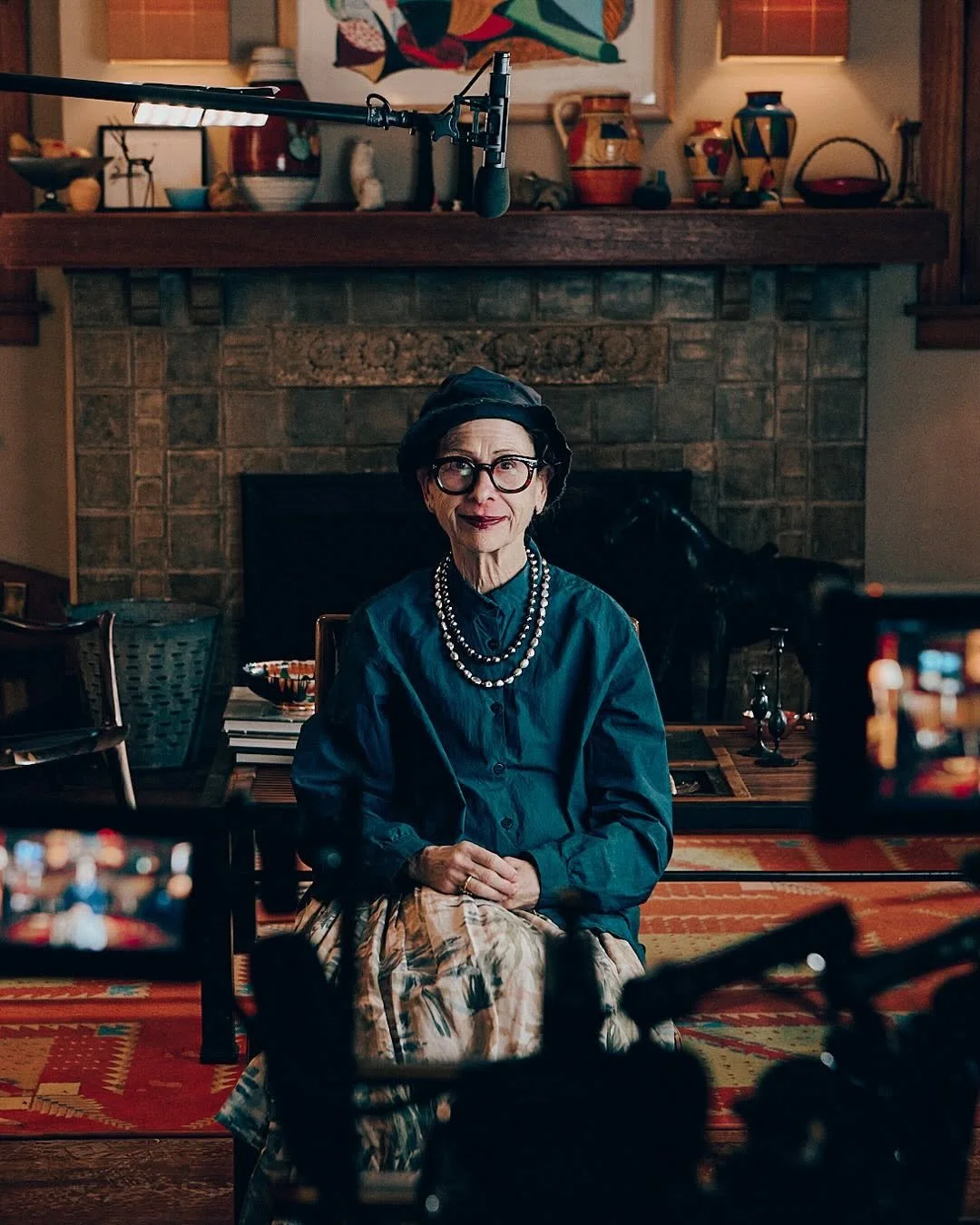 An elderly woman with glasses and dark hair, wearing a pearl necklace, a dark hat, a blue shirt, and a patterned skirt, sitting in front of a stone fireplace in a cozy room. There are multiple cameras and recording equipment set up around her, suggesting an interview or documentary scene.