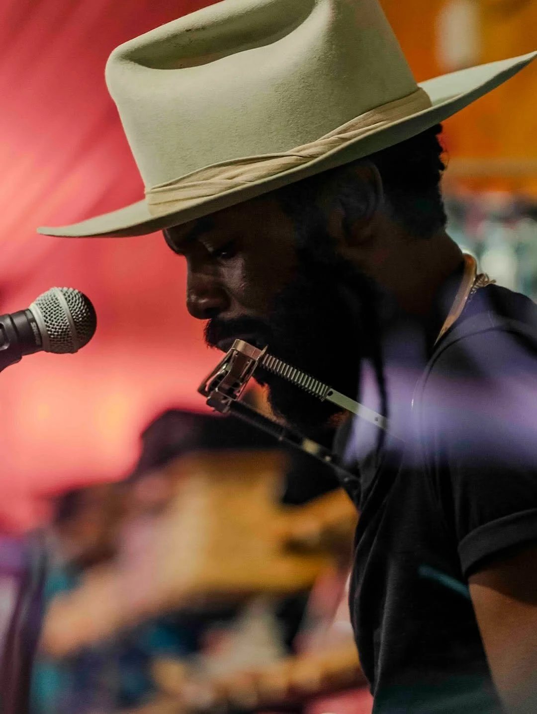 A man with a beard wearing a large beige hat and playing a harmonica in front of a microphone.