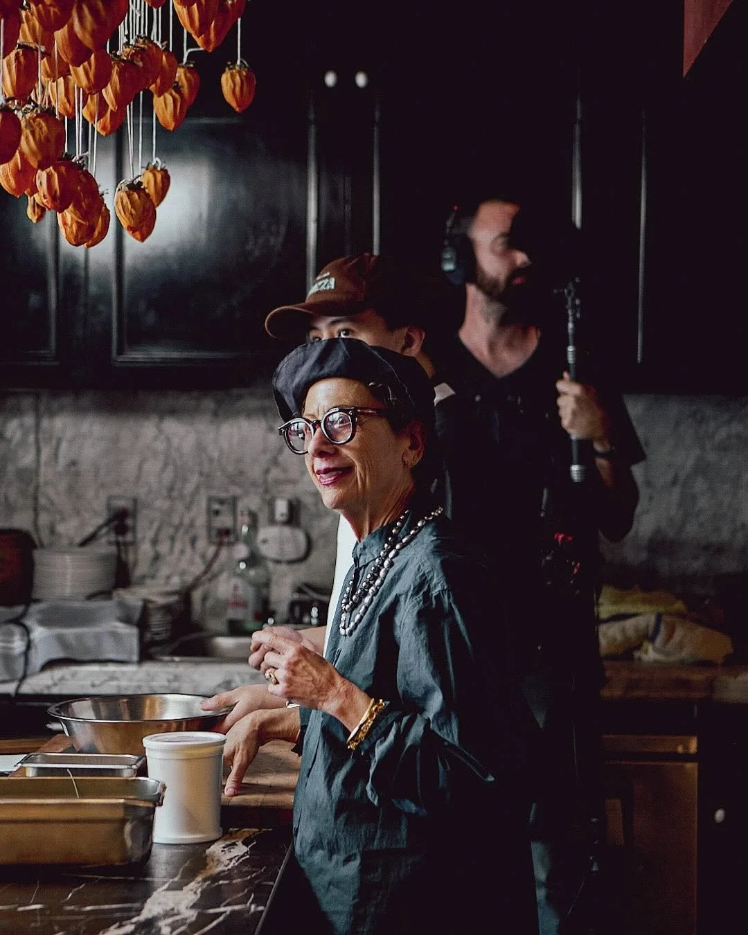 A woman in glasses and a black headscarf standing in a kitchen, smiling, with a man with headphones and a beard holding a boom microphone in the background. Hanging dried orange peppers are in the foreground.