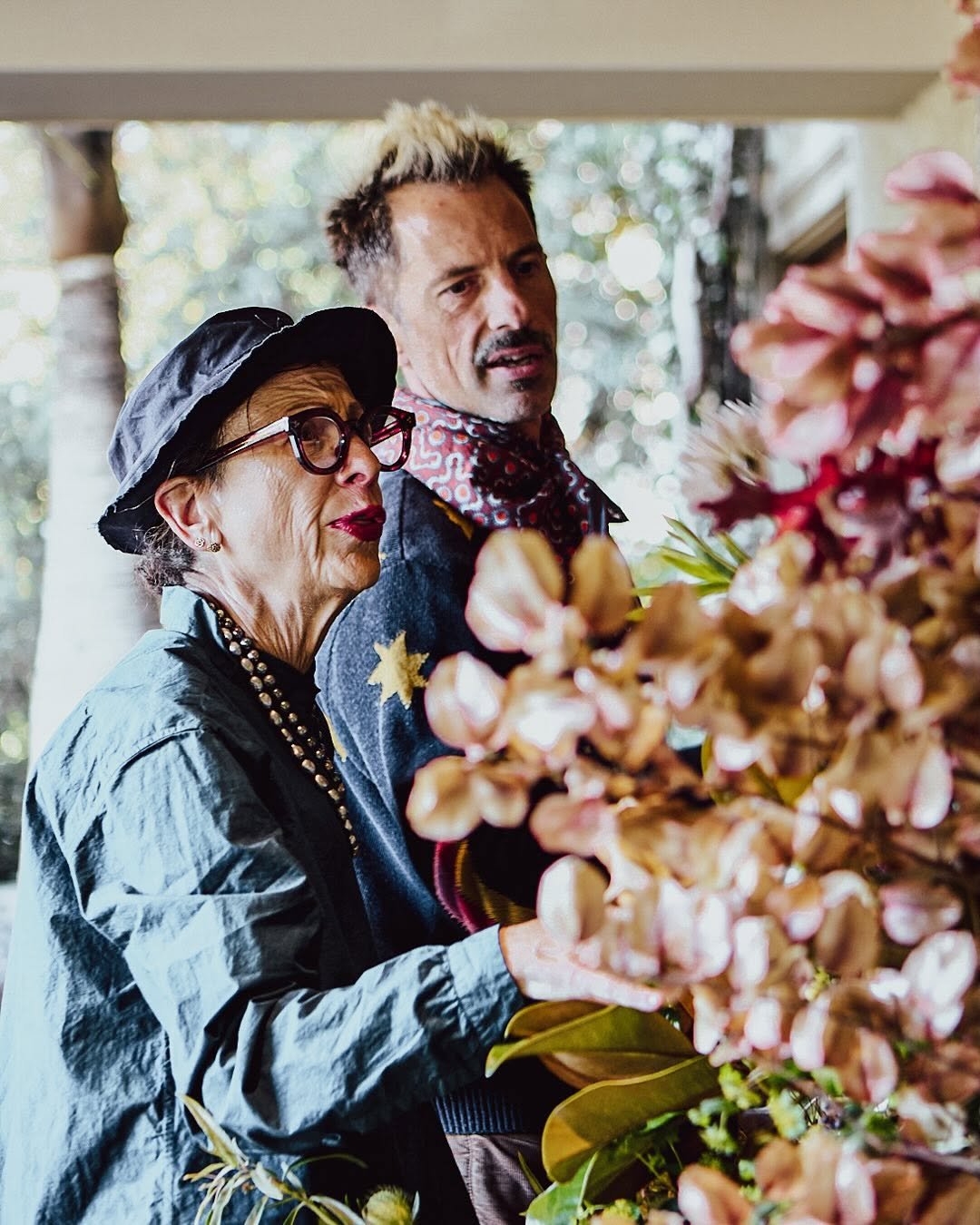 An older woman and a middle-aged man looking at a floral display, with the woman wearing a black hat, red glasses, and pearl necklace, and the man wearing a patterned scarf.